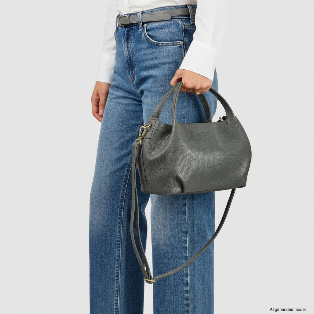 A person wearing a white shirt and blue jeans holds an Italian leather handbag with a top handle and detachable shoulder strap.