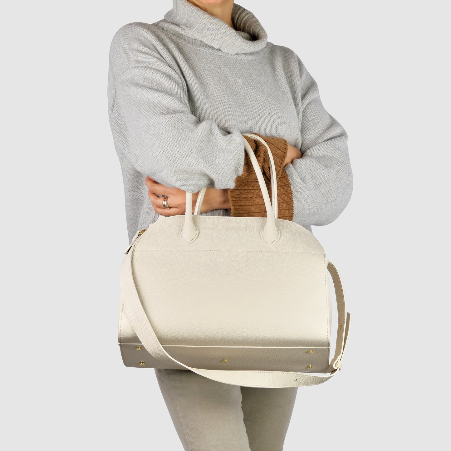 A person in a light gray turtleneck sweater holds a large Italian leather bag with gold studs and a shoulder strap, standing against a plain light background.