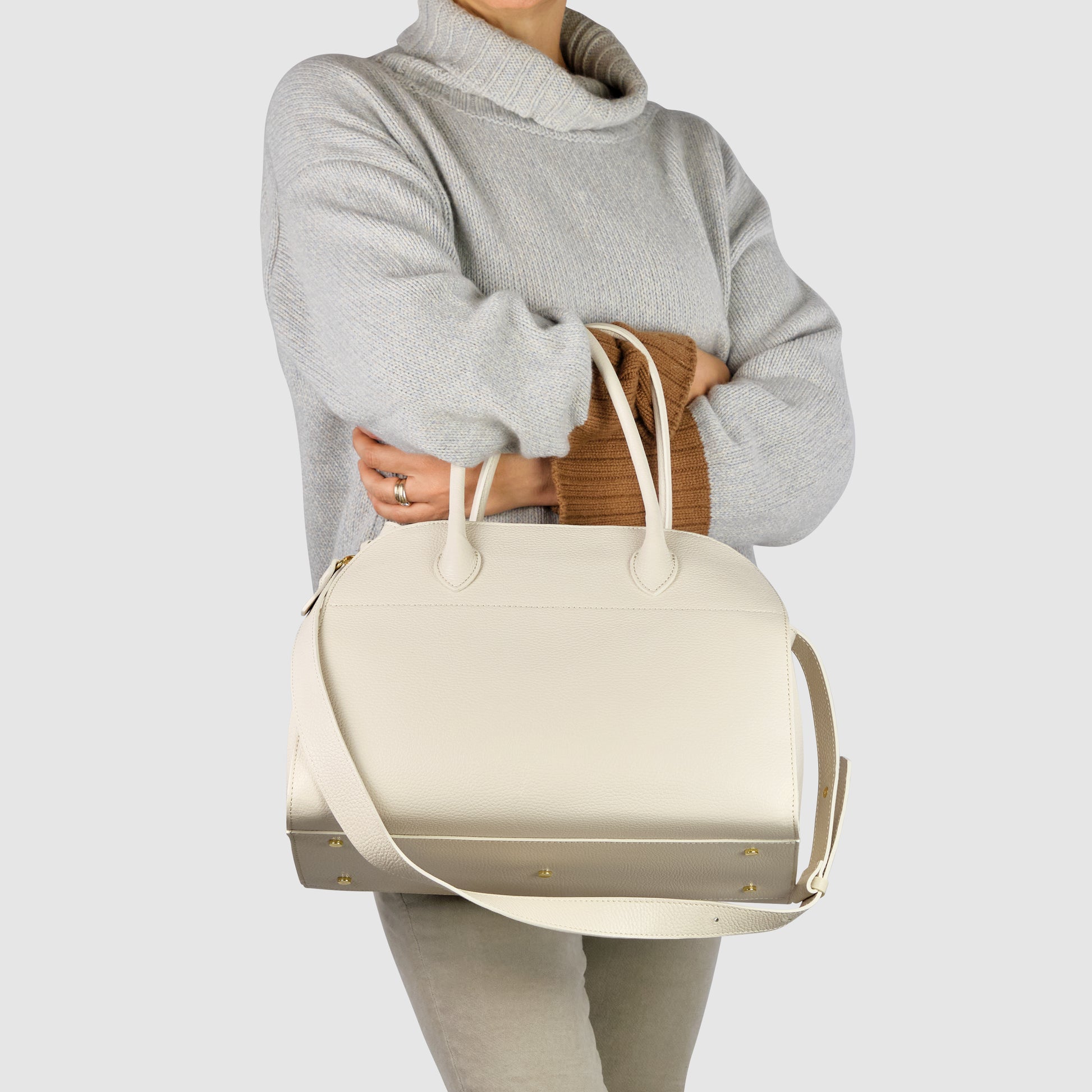 A person in a light gray turtleneck sweater holds a large Italian leather bag with gold studs and a shoulder strap, standing against a plain light background.