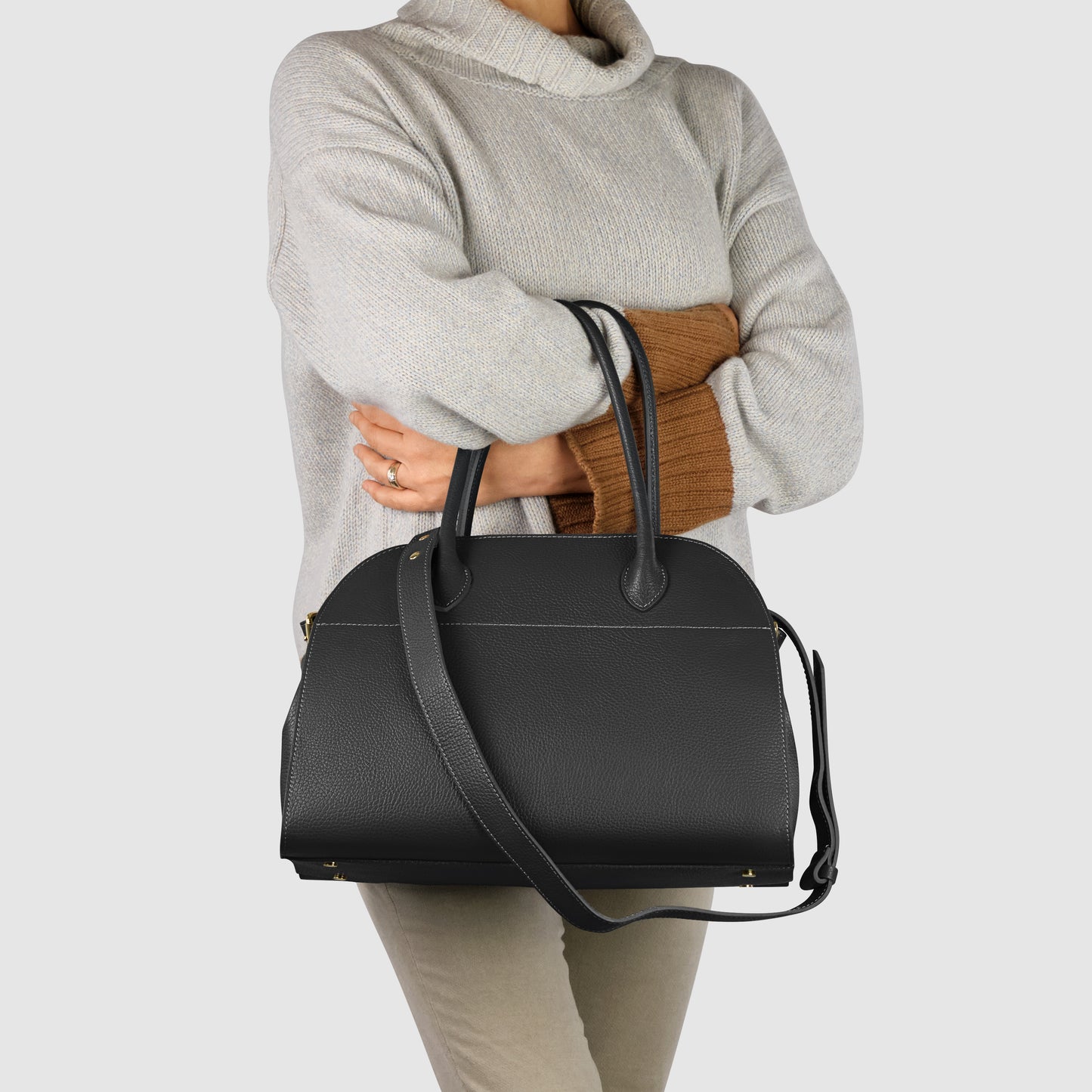 A person wearing a light gray turtleneck sweater and beige pants holds an Italian leather bag with top handles and a shoulder strap, standing against a plain light background.