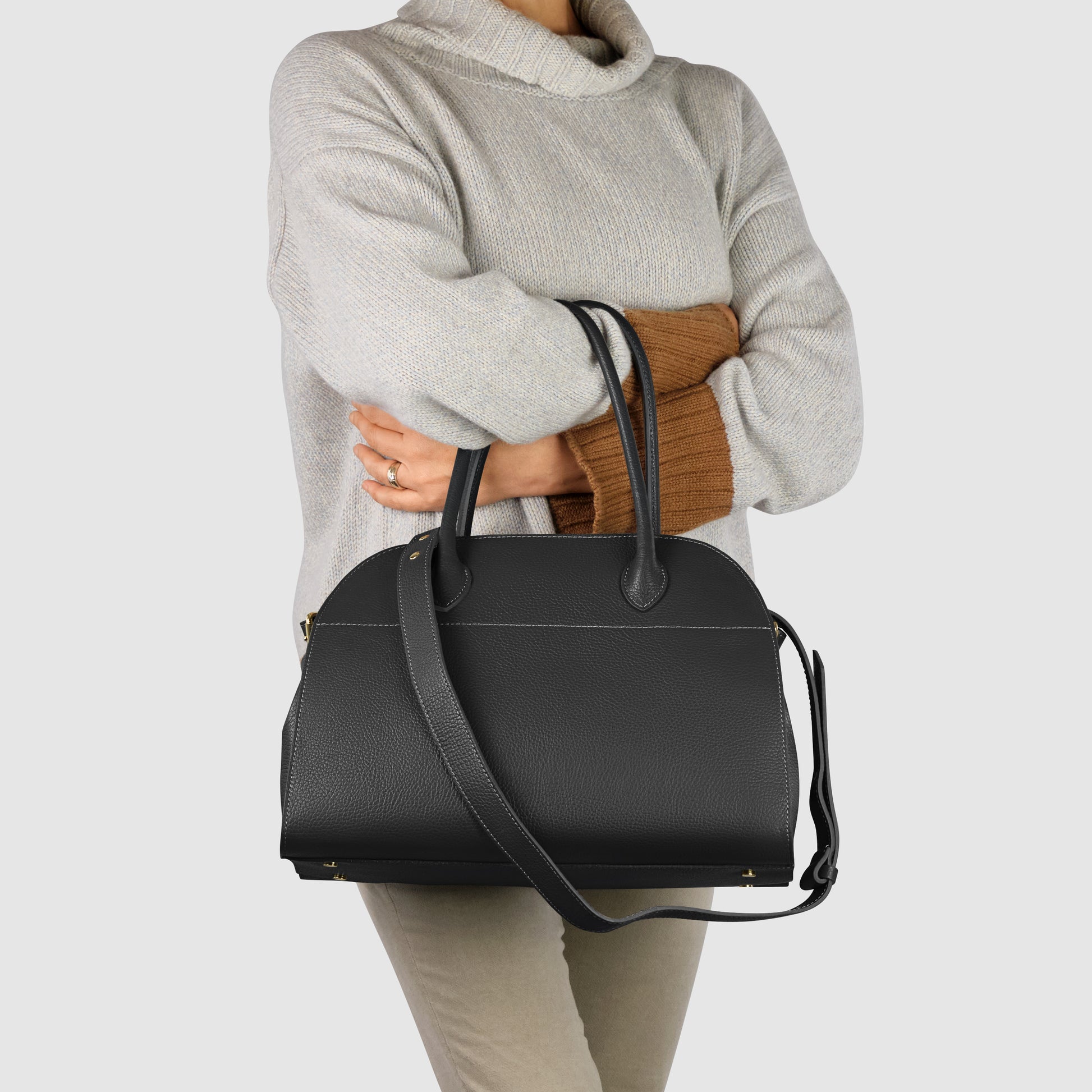 A person wearing a light gray turtleneck sweater and beige pants holds an Italian leather bag with top handles and a shoulder strap, standing against a plain light background.