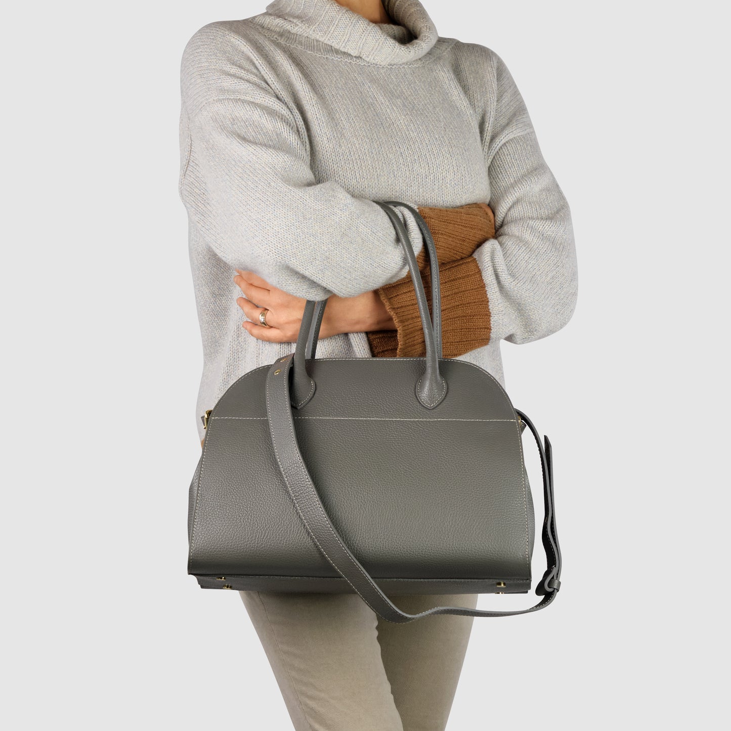 A person wearing a light gray turtleneck sweater and beige pants holds a large Italian leather bag with short handles and a long strap, against a plain light background.