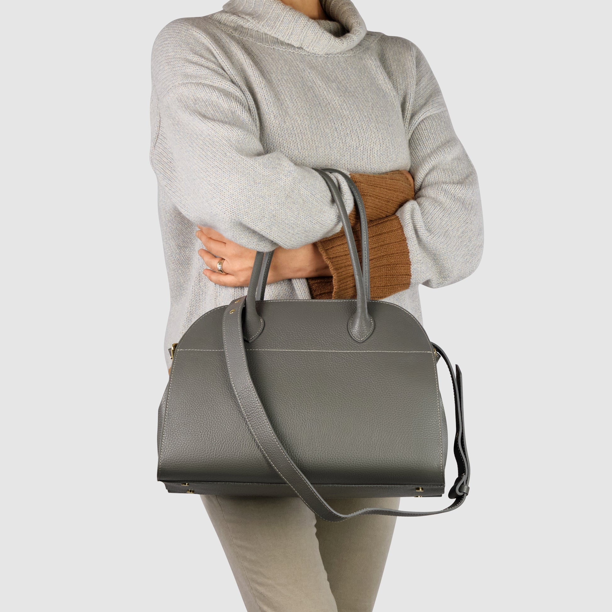 A person wearing a light gray turtleneck sweater and beige pants holds a large Italian leather bag with short handles and a long strap, against a plain light background.