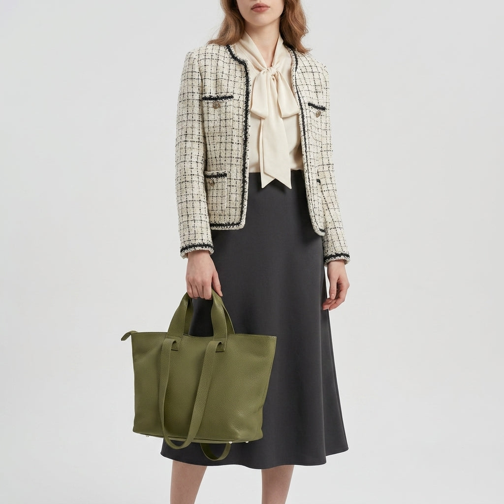 A woman wearing a cream plaid jacket, light blouse with a bow, and dark gray skirt stands holding a large trapezoid tote bag. The background is plain white. Her face is partially out of frame.