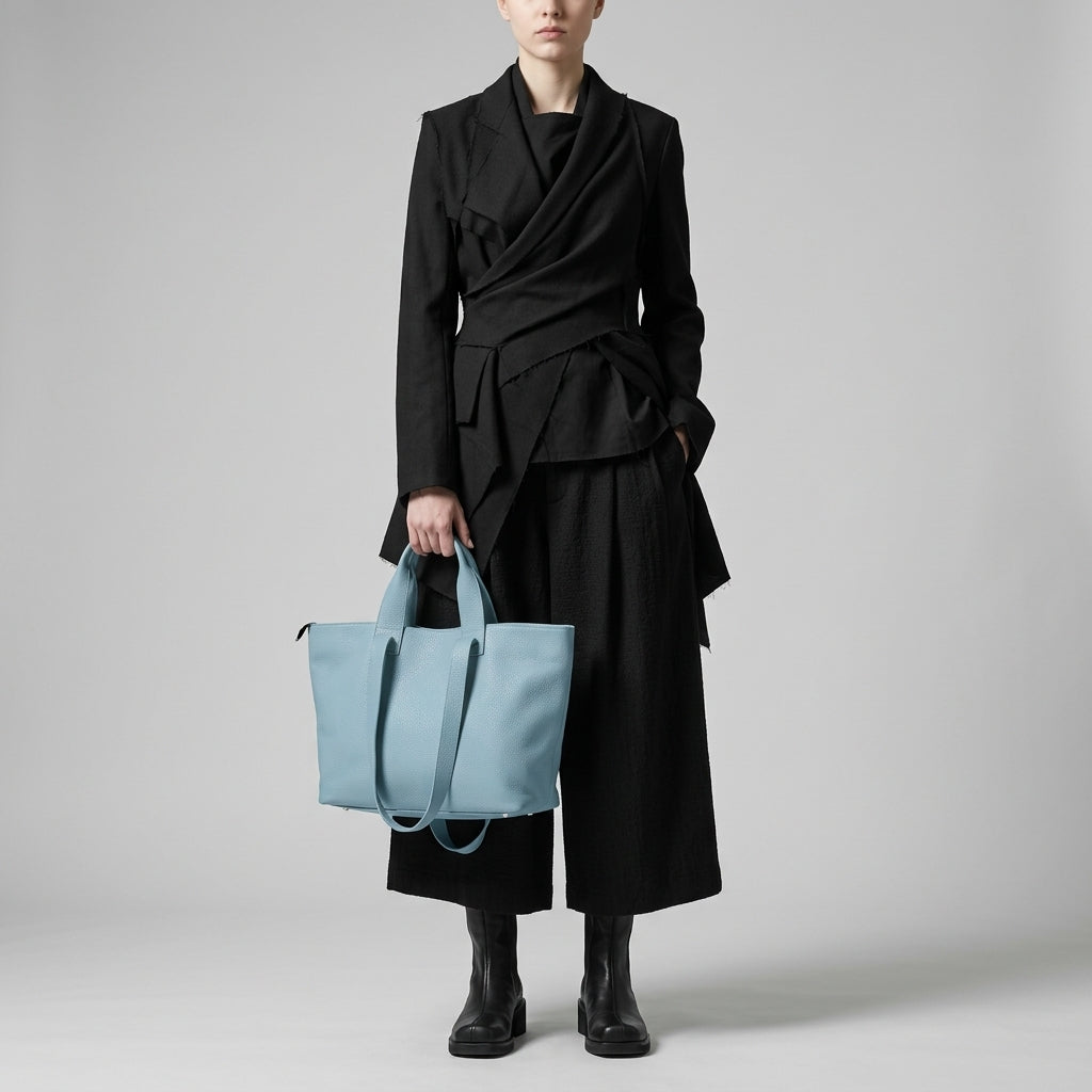 A person wearing a stylish black blazer, wide-leg black pants, and black boots stands holding a large light blue trapezoid tote bag against a plain light gray background. Their face is not fully visible.