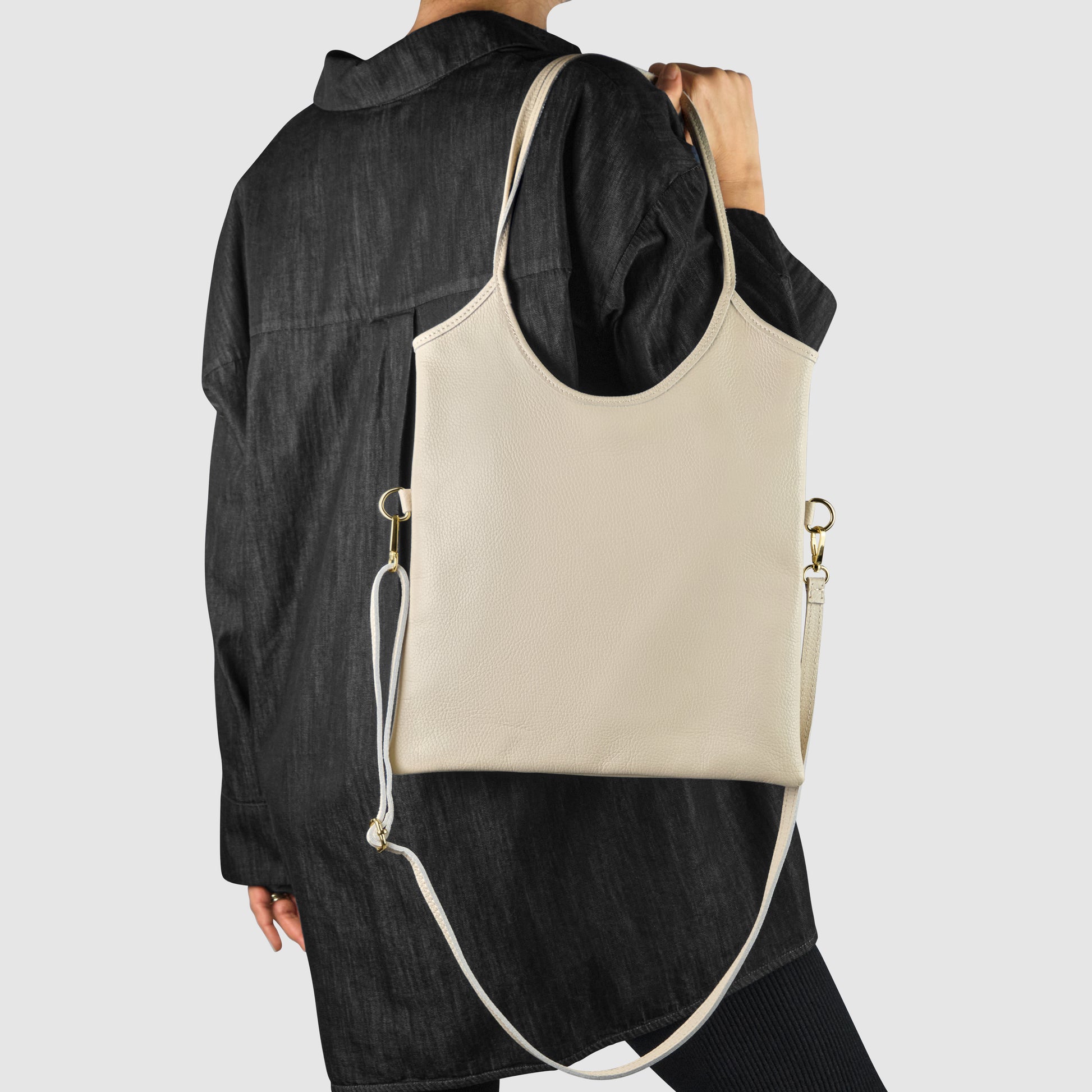 A person wearing a loose black jacket holds an Italian leather everyday shoulder bag with gold hardware and an adjustable strap, showcasing Florentine craftsmanship, over their shoulder as they face away from the camera against a plain background.