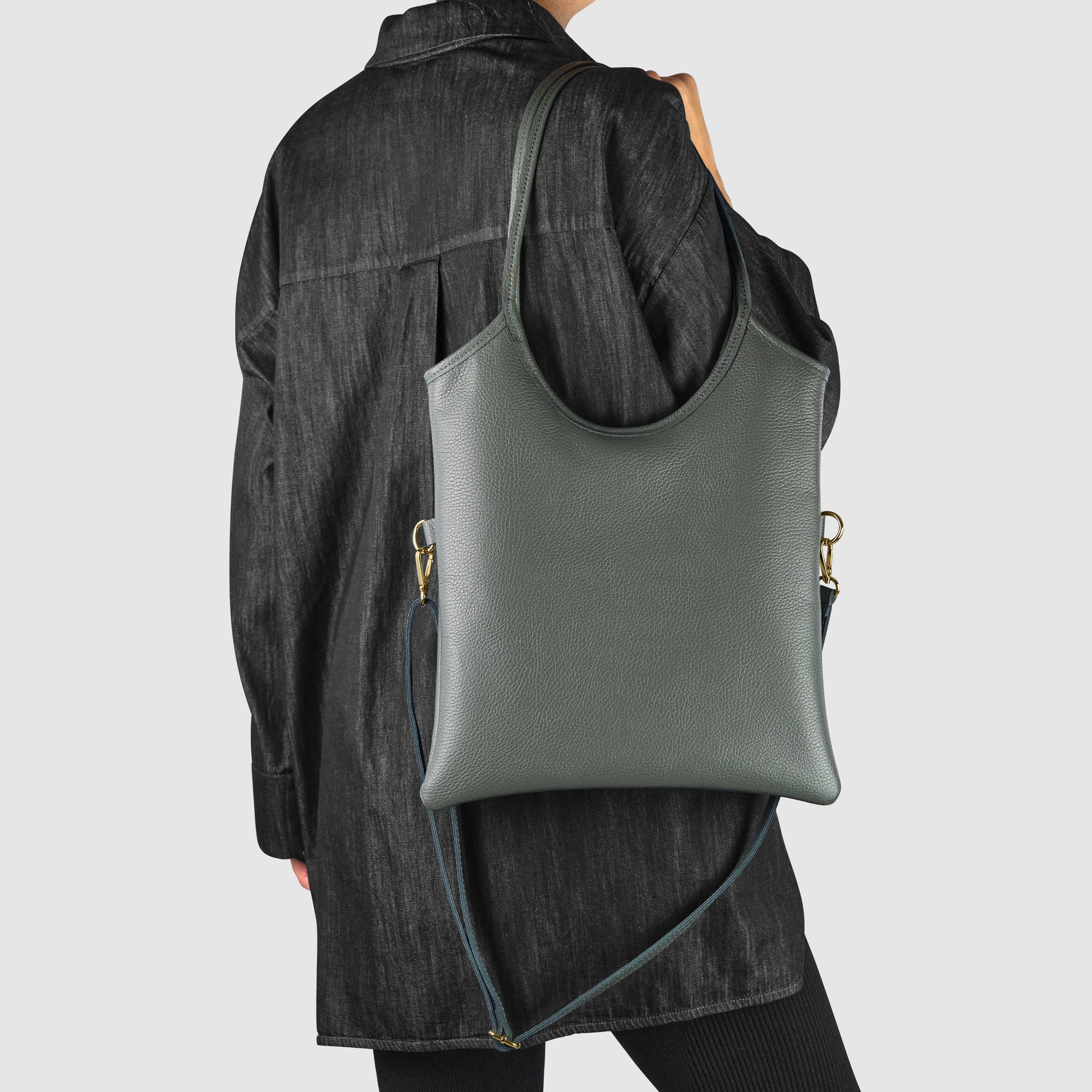 A person wearing a loose, black denim jacket and black pants is holding an everyday shoulder bag with a detachable strap, crafted with Florentine craftsmanship, over their shoulder, facing away from the camera.