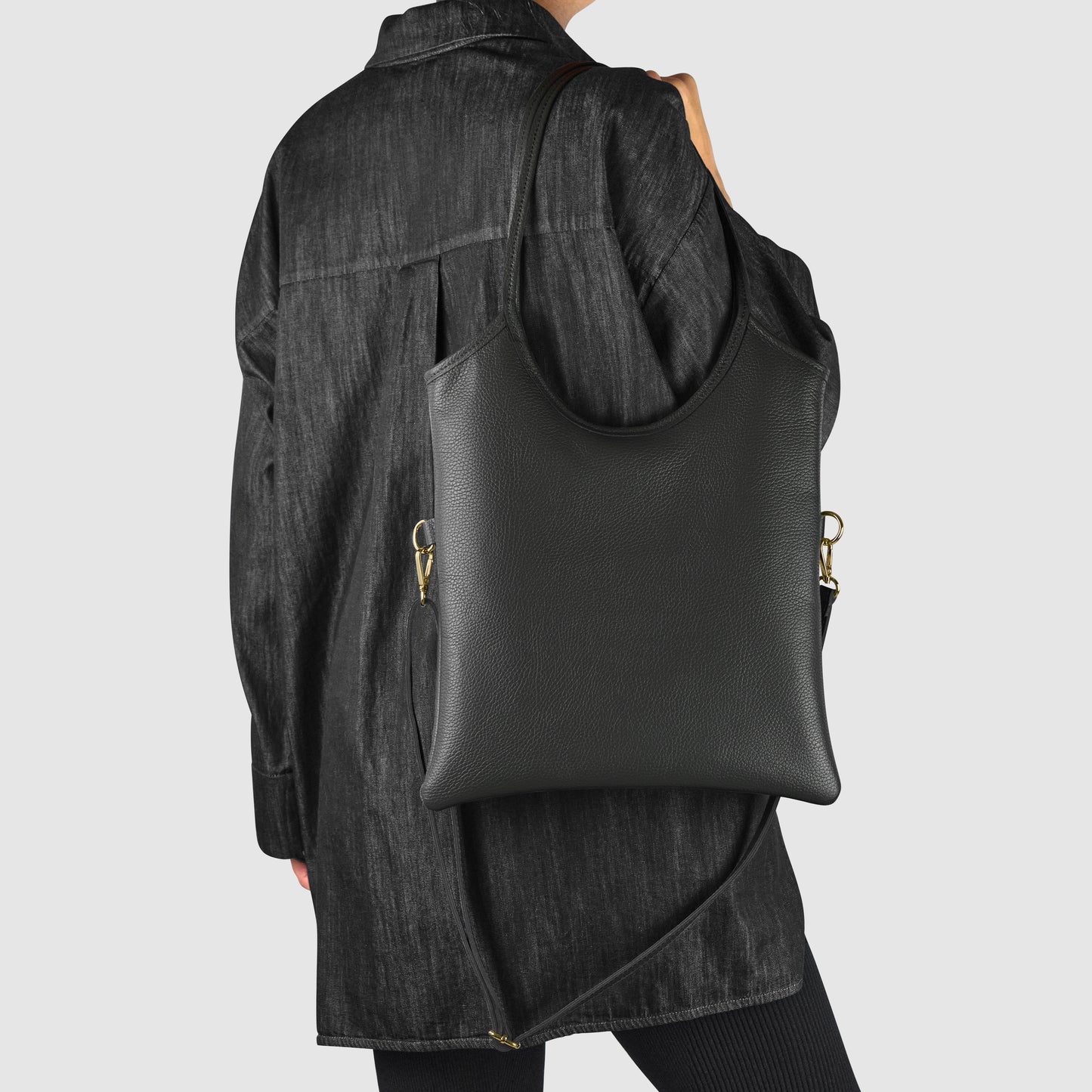 A person wearing a long, dark textured coat carries an Italian leather bag with gold hardware over their shoulder. The background is plain and light-colored.