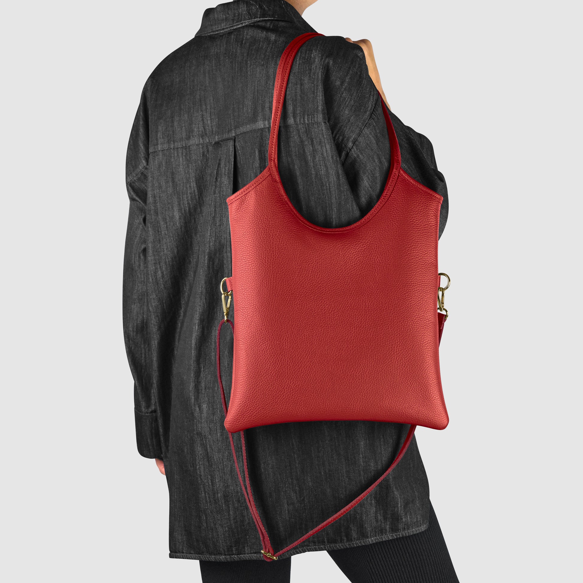A person in a loose black jacket and pants carries a large red Italian leather bag with a detachable shoulder strap, shown from the back against a plain light background—a striking example of Florentine craftsmanship.