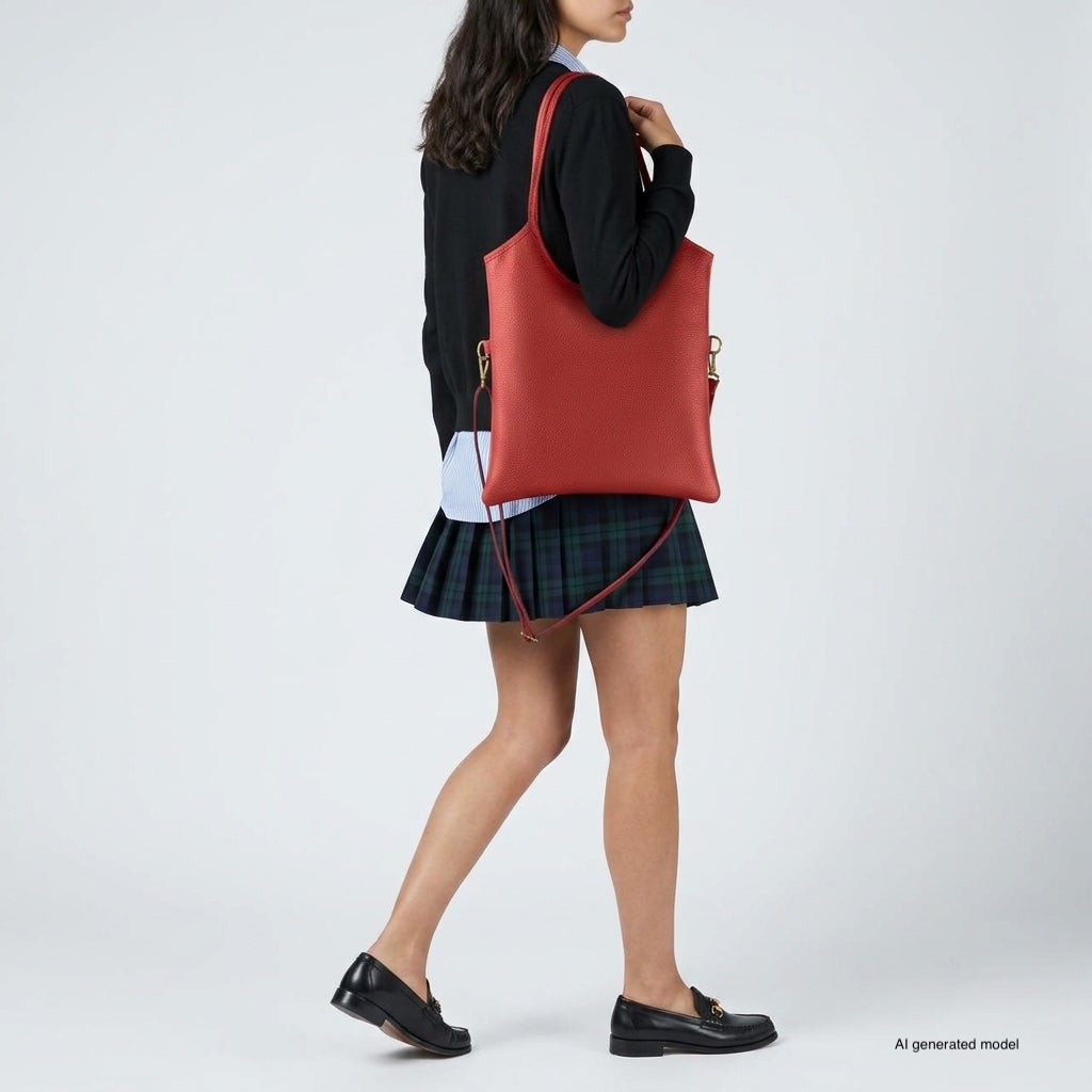 A woman wearing a black sweater, plaid skirt, and black loafers carries an elegant Italian leather tote over her shoulder, standing against a plain white background.