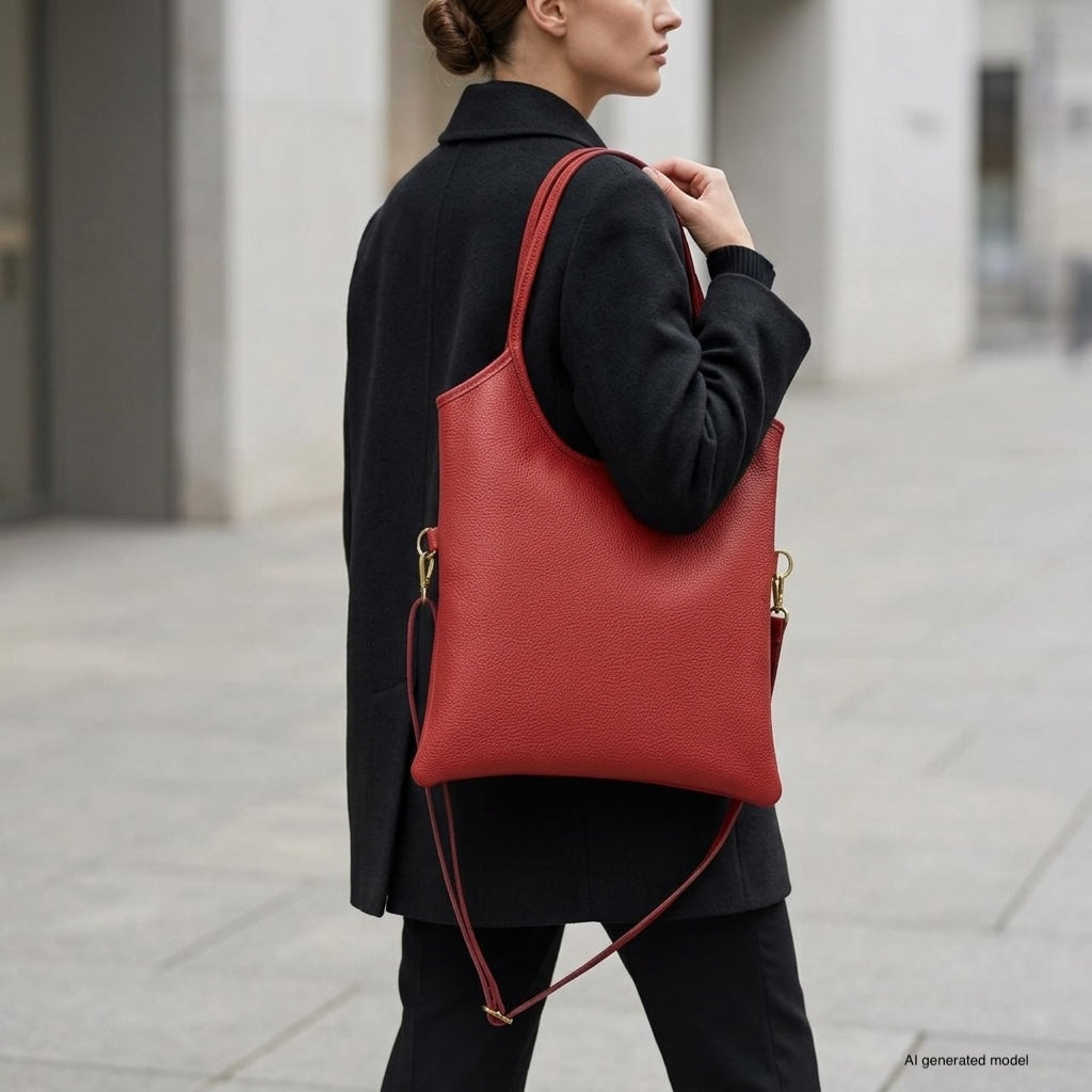 A woman in a black coat walks outdoors, carrying a large Italian leather tote over her shoulder. The urban background features gray pavement and light-colored buildings.