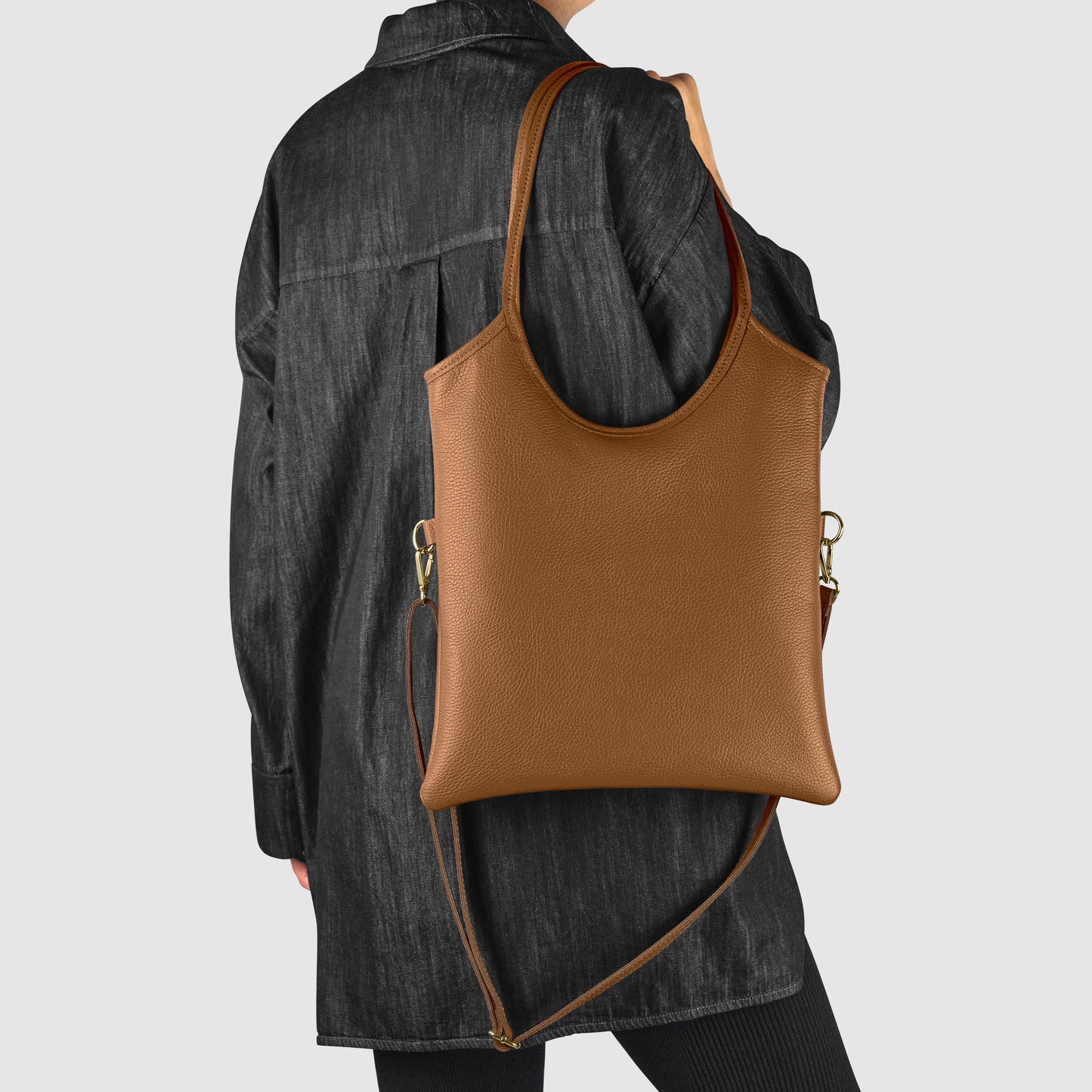 A person wearing a loose, black jacket and black pants is shown from the back, carrying a large Italian leather bag with gold hardware. The background is plain light gray, highlighting the elegance of this everyday shoulder bag.
