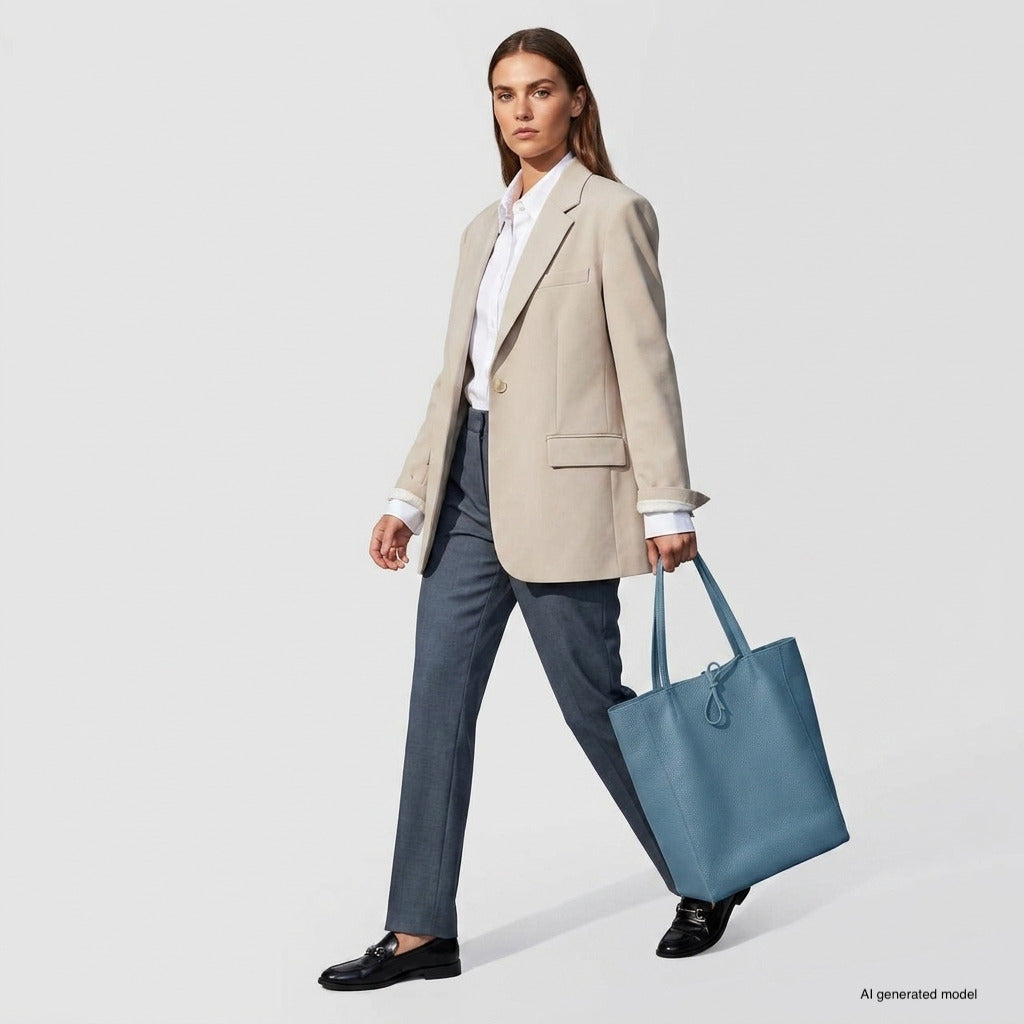 A woman wearing a beige blazer, white shirt, gray pants, and black loafers carries a large women's carryall tote against a plain light background.