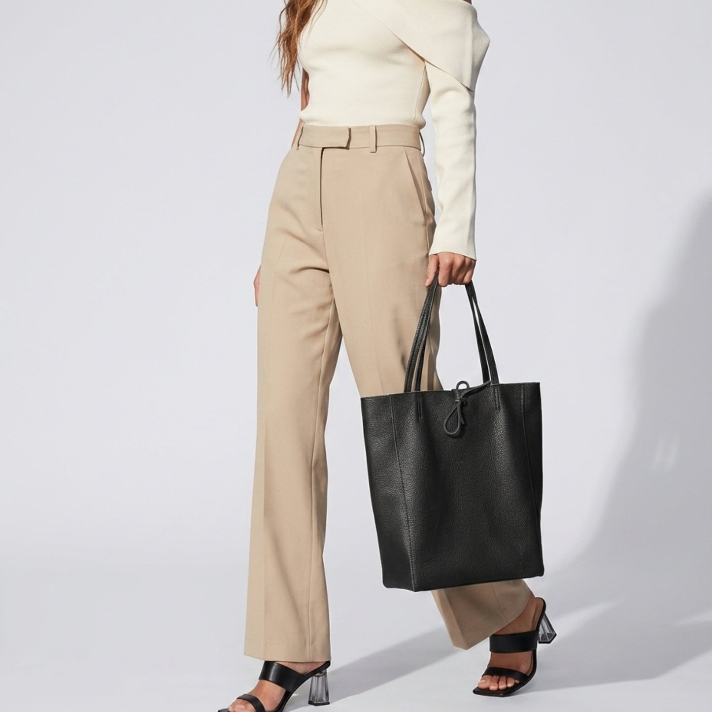 A woman in a cream off-the-shoulder top and beige high-waisted pants holds an Italian leather tote—a sleek women’s everyday carryall. She pairs it with black heeled sandals against a plain, light-colored backdrop.