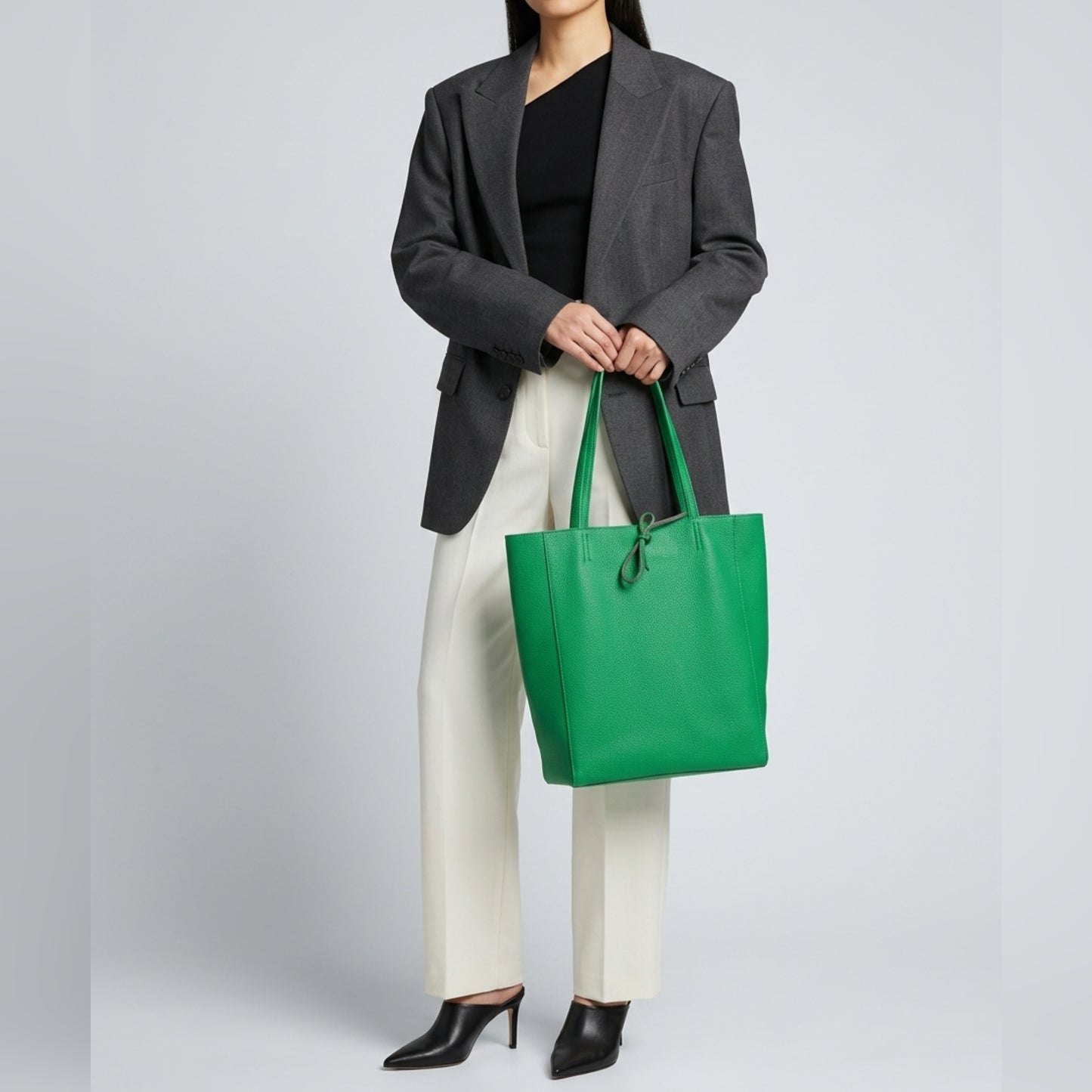 A woman stands against a plain backdrop in a gray blazer, black top, and white pants, holding a large green Italian leather tote by her side. Her black heeled shoes complete the look. Her face is not visible.