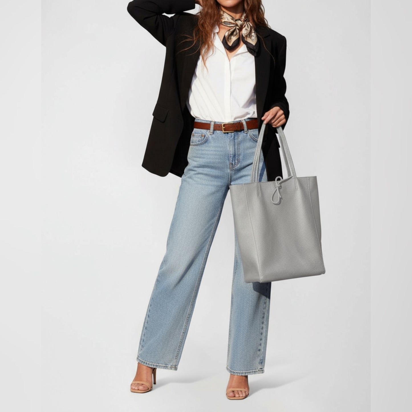 A woman wearing a black blazer, white shirt, blue jeans, and heels poses with an Italian leather tote and a patterned scarf around her neck. Her hand is raised to her head, and her face is partially out of frame.