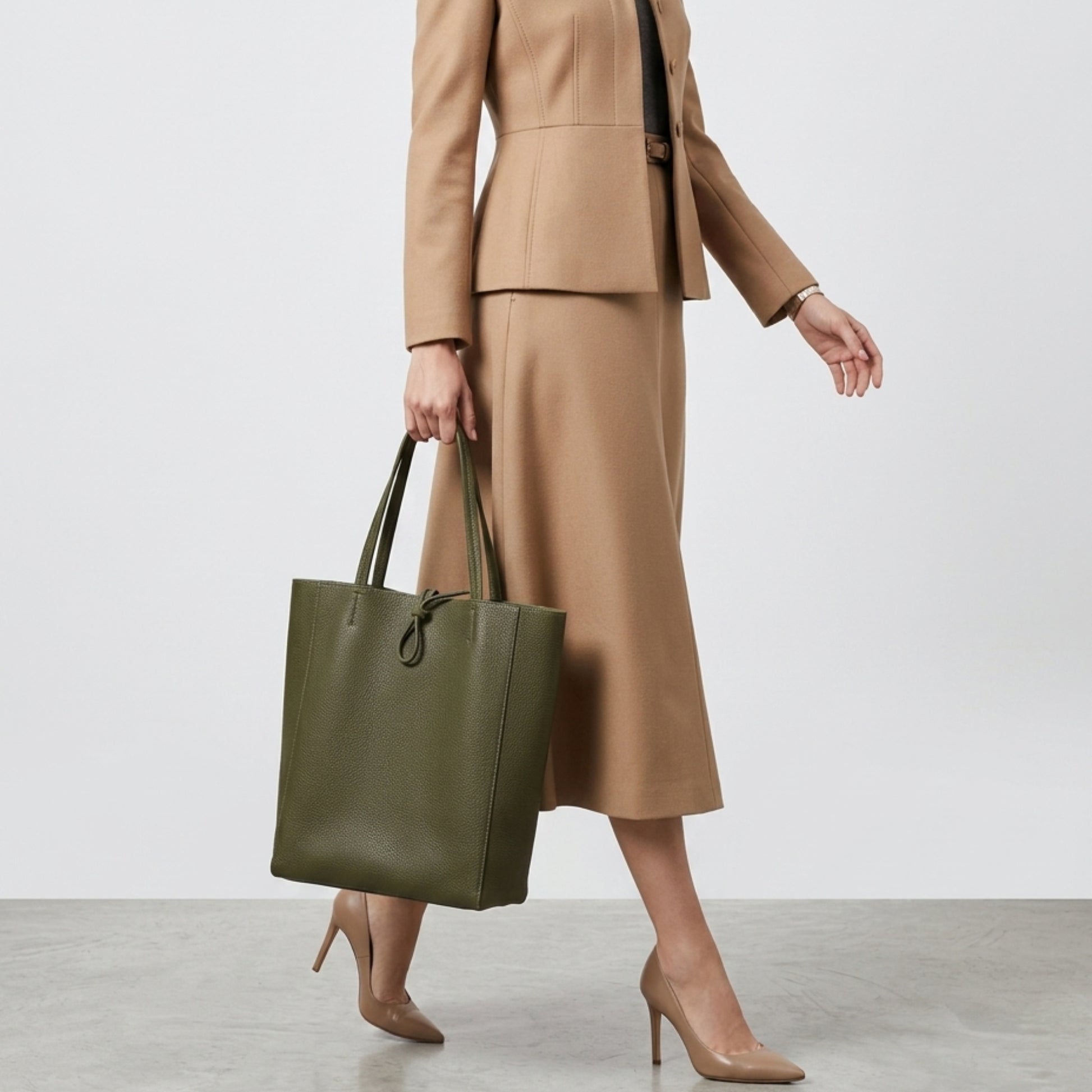 A woman wearing a tan blazer, matching midi skirt, and nude heels walks while carrying an Italian leather tote in olive green—a stylish women's everyday carryall. The background is simple and neutral.