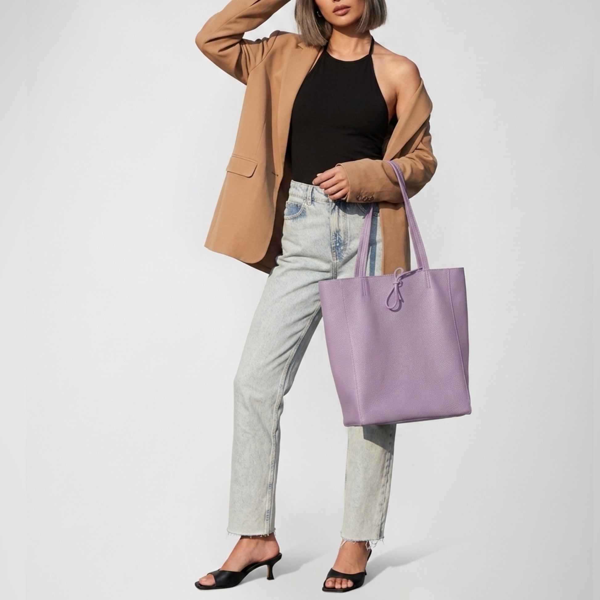 A woman in a tan blazer, black halter top, light-wash jeans, and black heeled sandals holds a women's everyday carryall—the large lavender tote—while posing with one hand on her head against a plain light background.