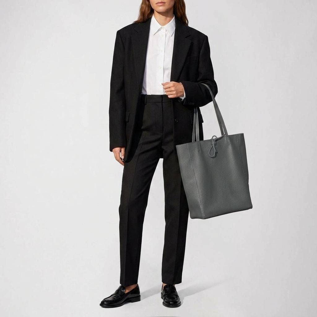 A person dressed in a black suit, white button-up shirt, and black loafers stands holding an Italian leather tote. The large pebbled calfskin bag adds elegance against the plain, light-colored background.