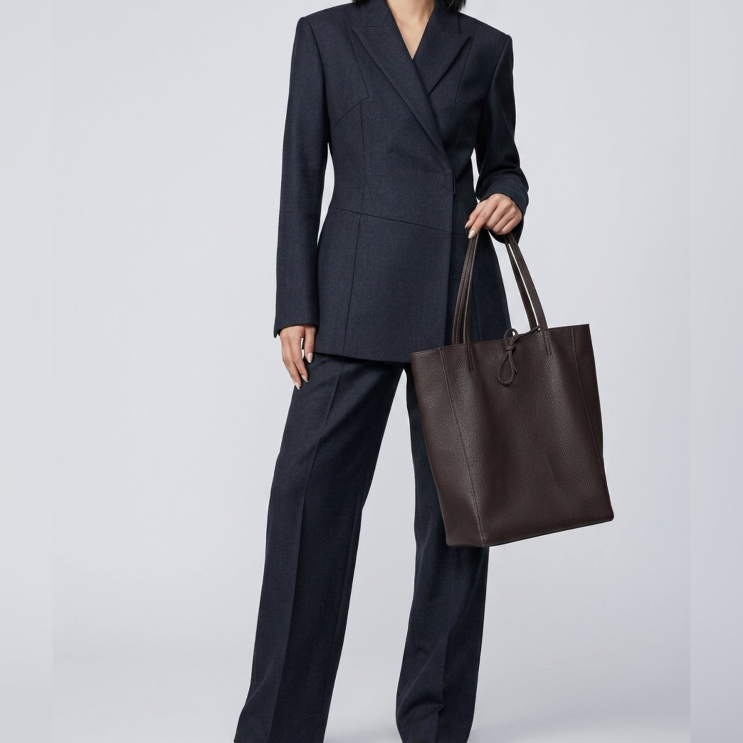 A person wearing a dark tailored pantsuit holds an Italian leather tote by the handles. The large brown women's everyday carryall stands out against the plain, light-colored background. The person's head is not visible in the image.