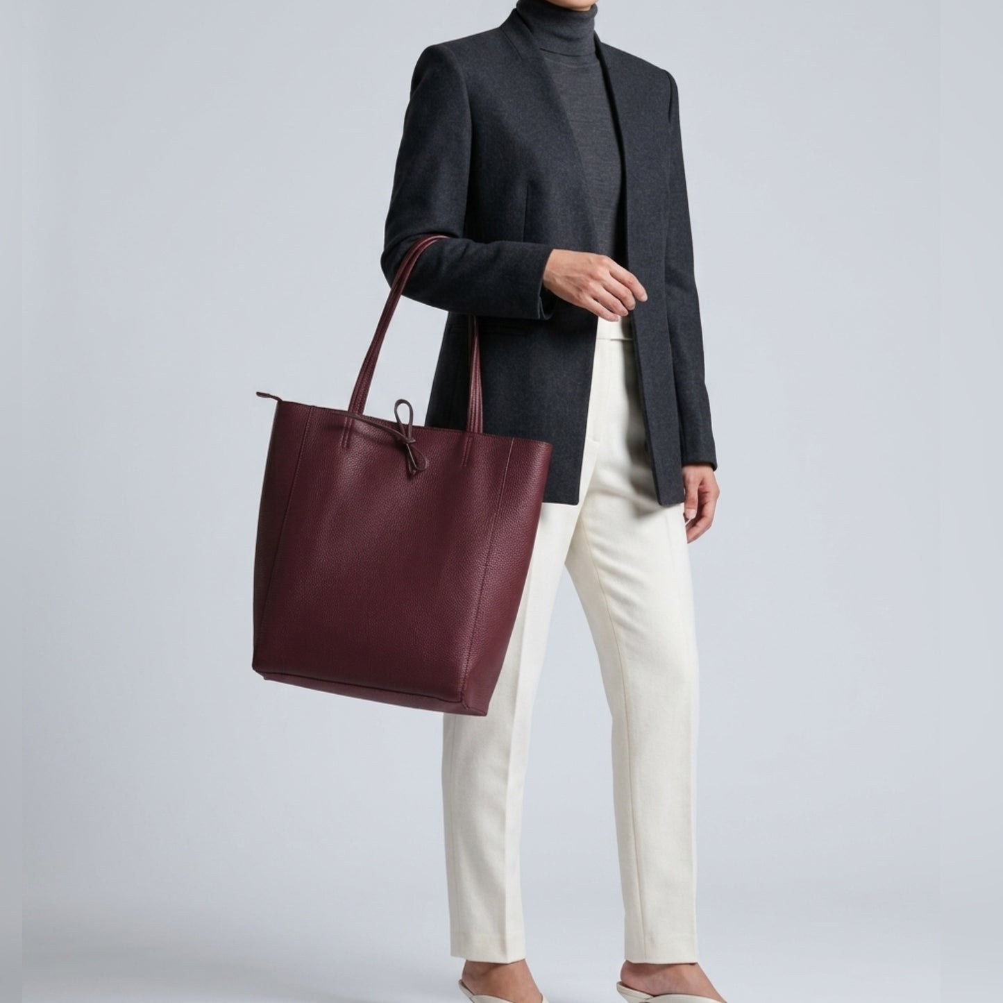 A person in a dark blazer, gray turtleneck, and white pants holds an Italian leather tote—crafted from pebbled calfskin—against a plain light gray background. They complete the look with beige flats.