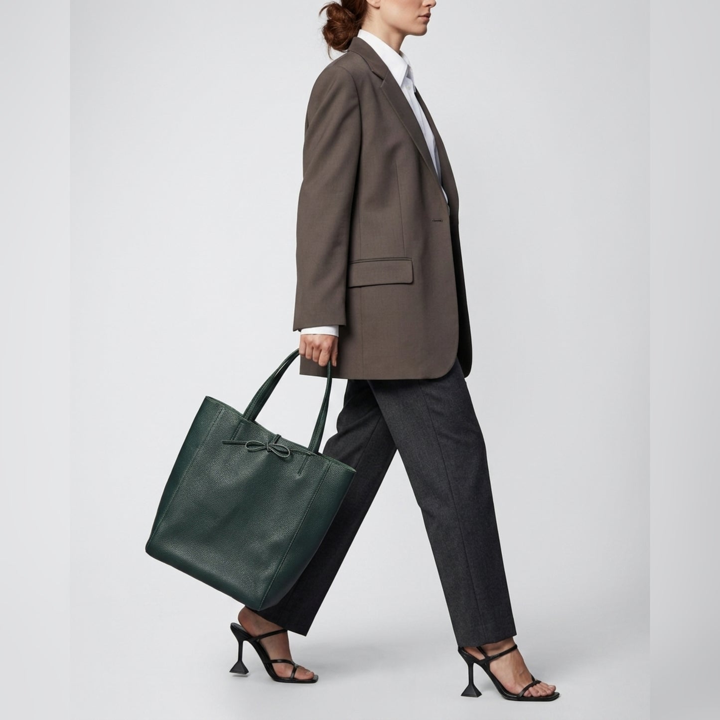 A woman in a brown blazer, white shirt, and dark trousers walks in black heeled sandals, carrying a large Italian leather tote with a bow detail—an ideal women’s everyday carryall—against a plain light background.