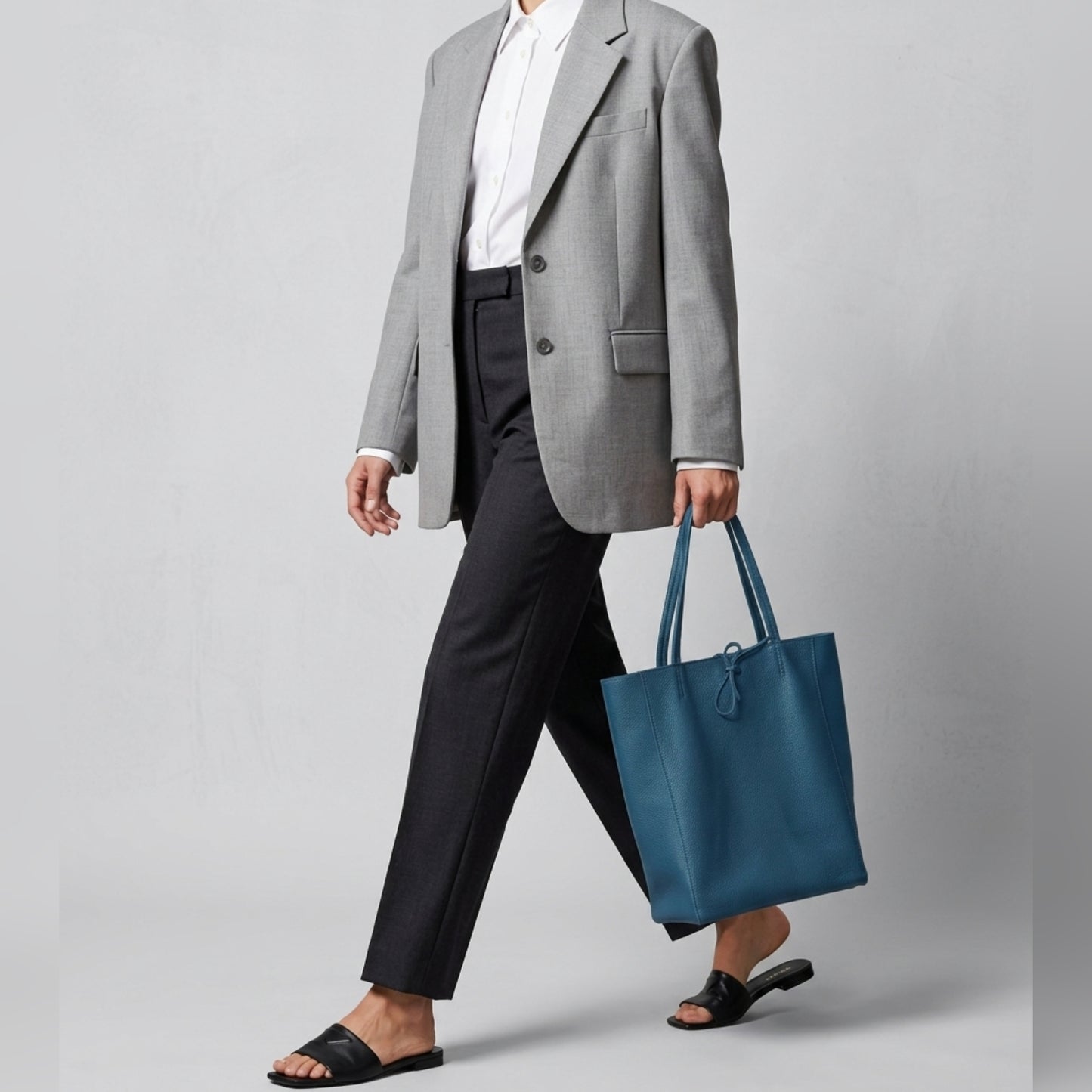 A person dressed in a gray blazer, white button-up shirt, black pants, and black sandals walks while carrying a pebbled calfskin bag against a plain light background.