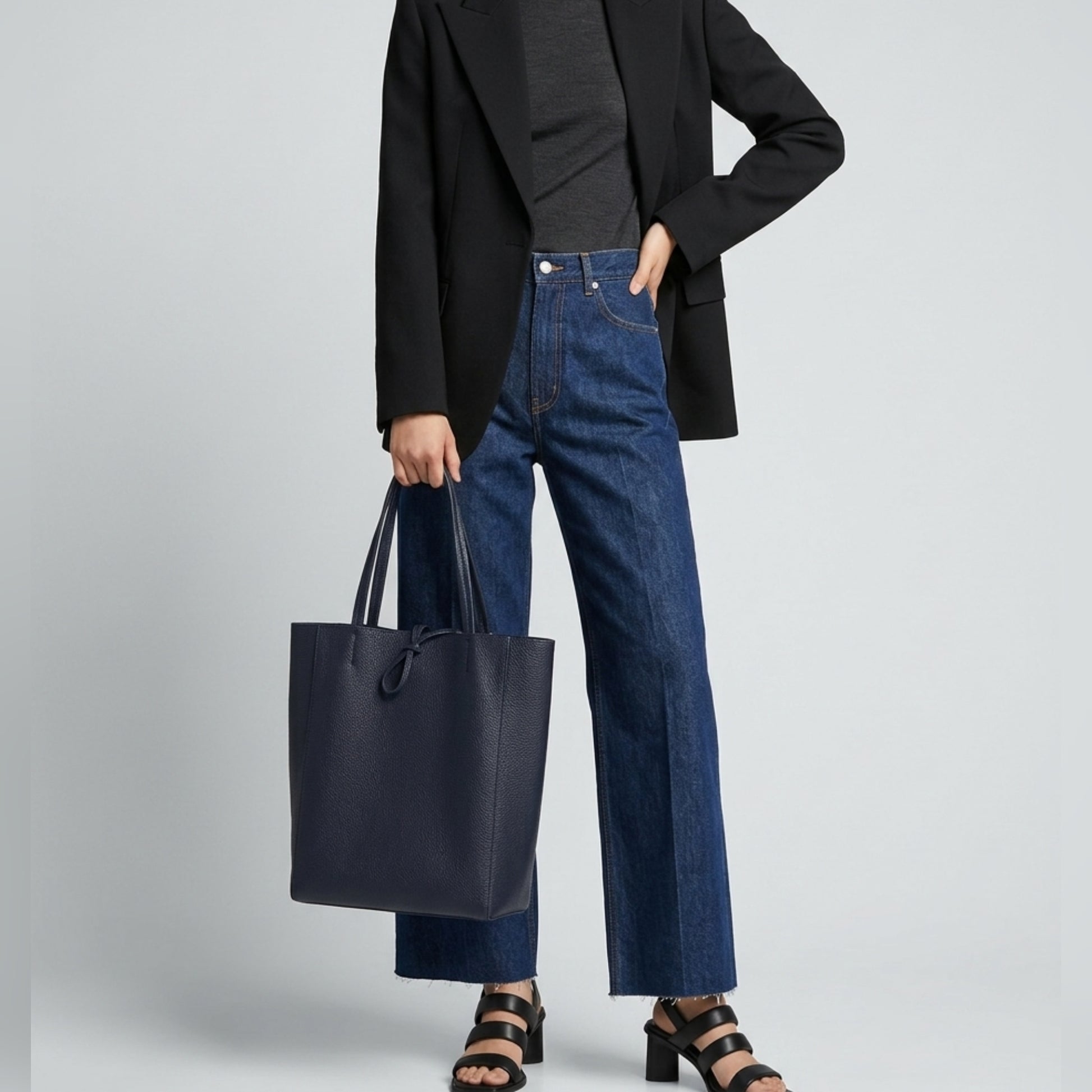 Person wearing a black blazer, dark top, blue high-waisted jeans, and black sandals holds an Italian leather tote—a chic women's everyday carryall—against a plain light background. Their face is not visible.
