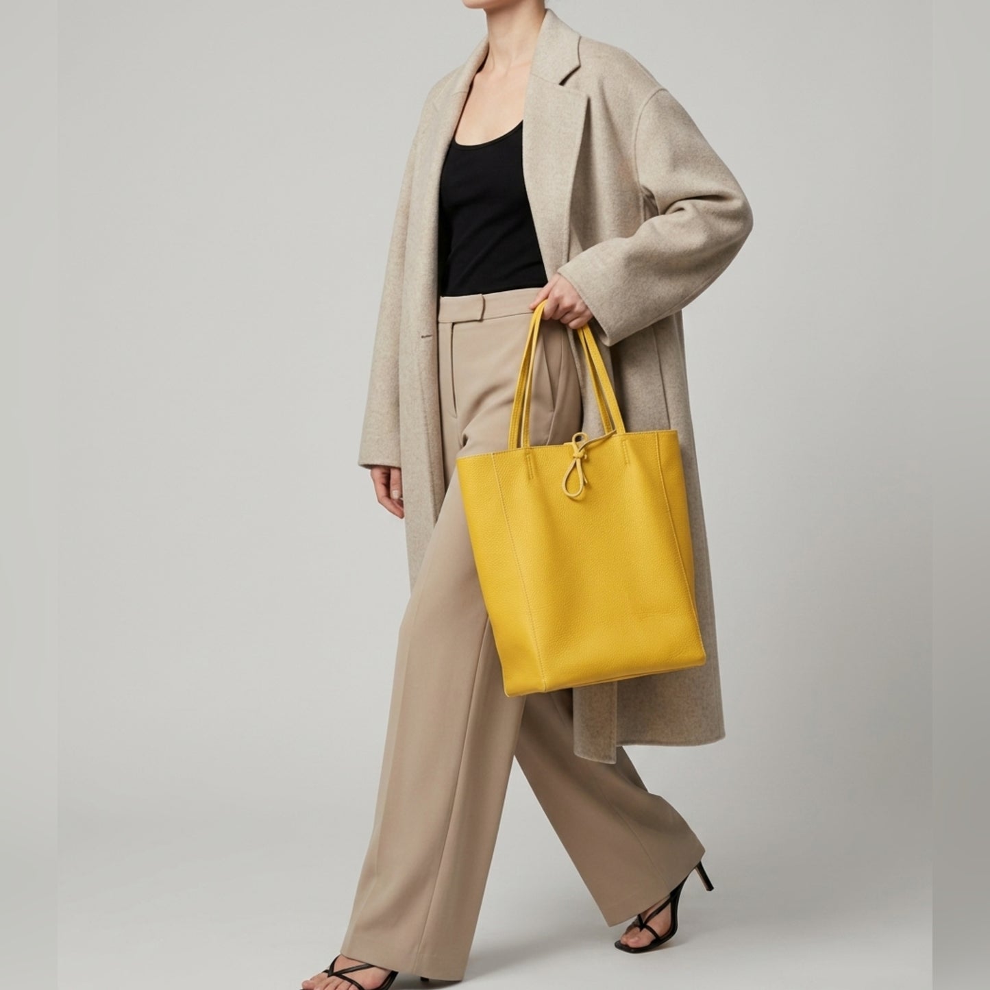 A woman wearing a beige coat, black top, and beige trousers carries an Italian leather tote in yellow. She is walking in black heeled sandals against a plain background.
