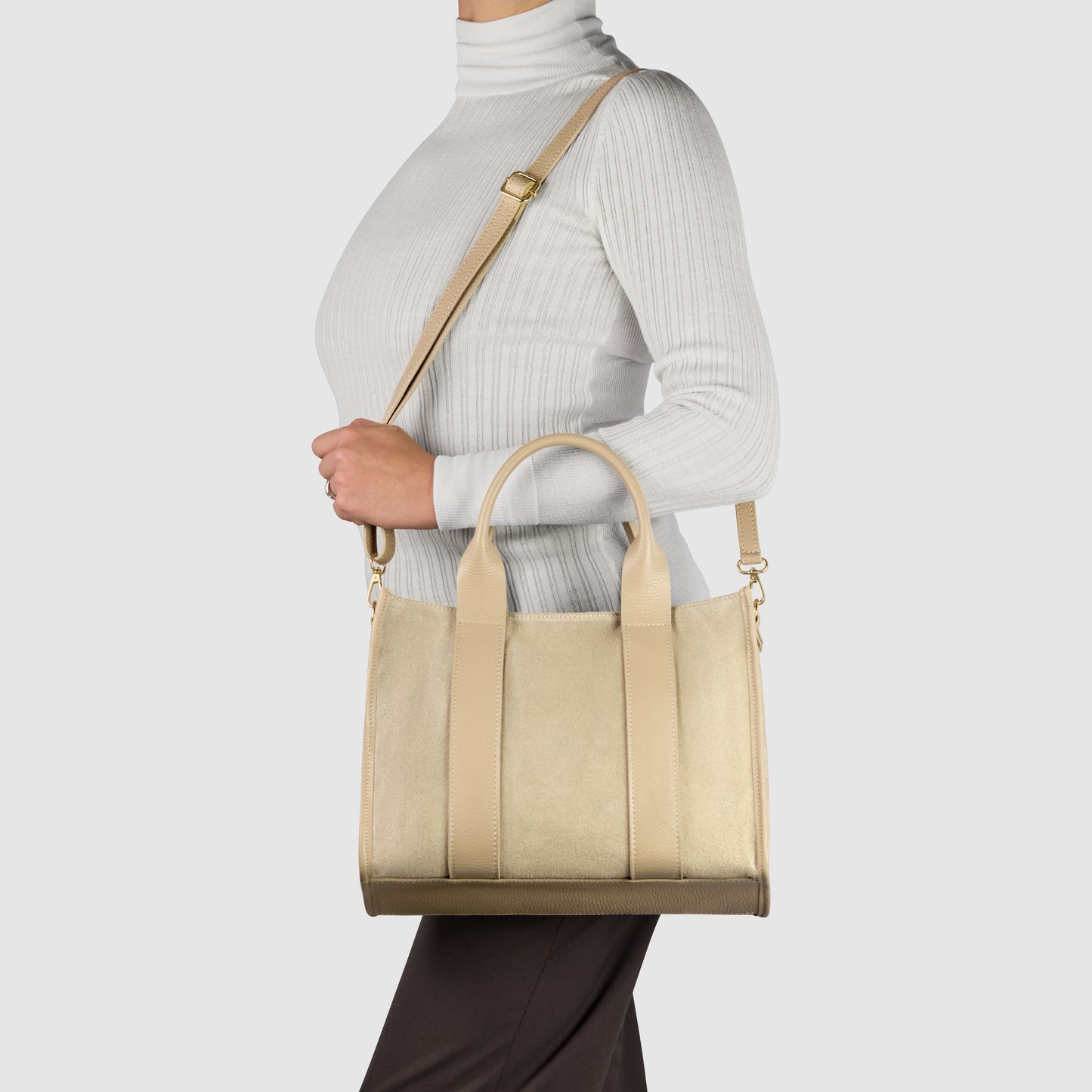 A person wearing a white ribbed turtleneck and black pants holds a handmade Italian bag with short handles and a long shoulder strap, styled as a crossbody shoulder bag, against a plain light background.