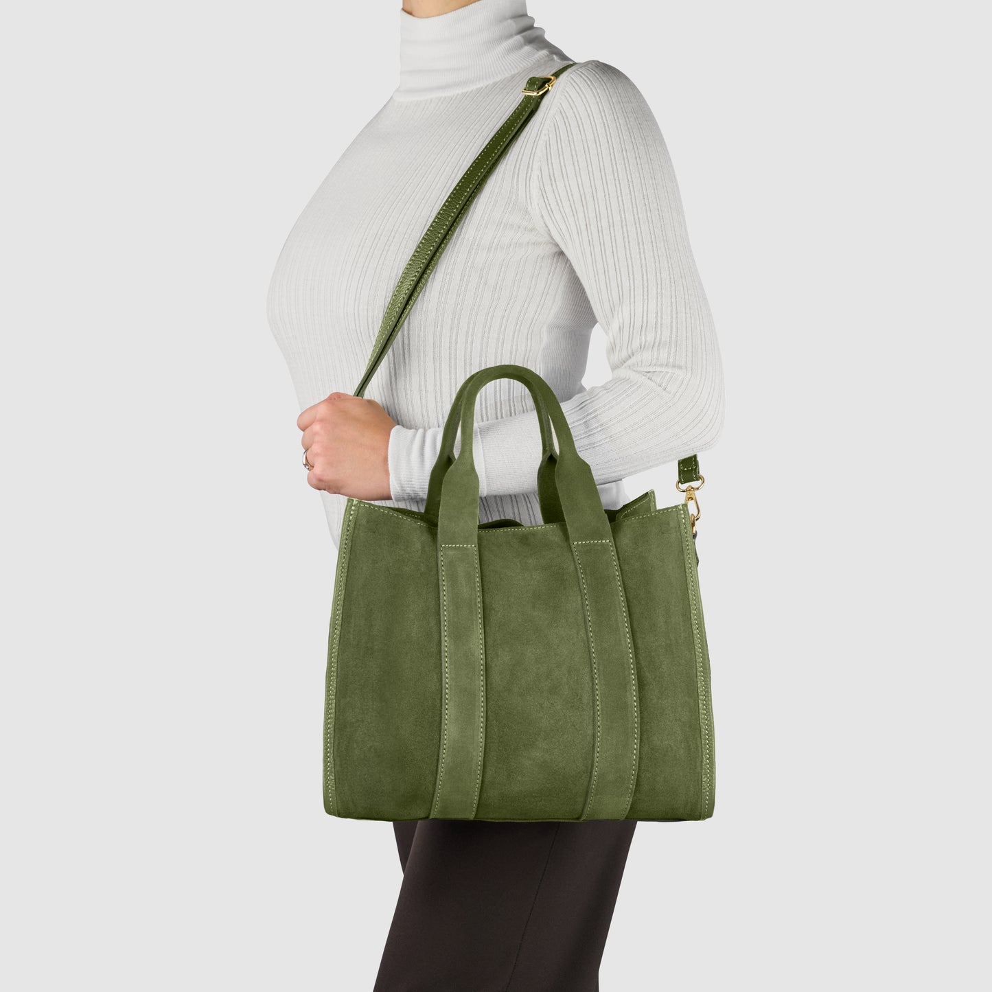 A person wearing a white turtleneck and black pants carries a green suede carryall with both handles and a shoulder strap against a plain light background.
