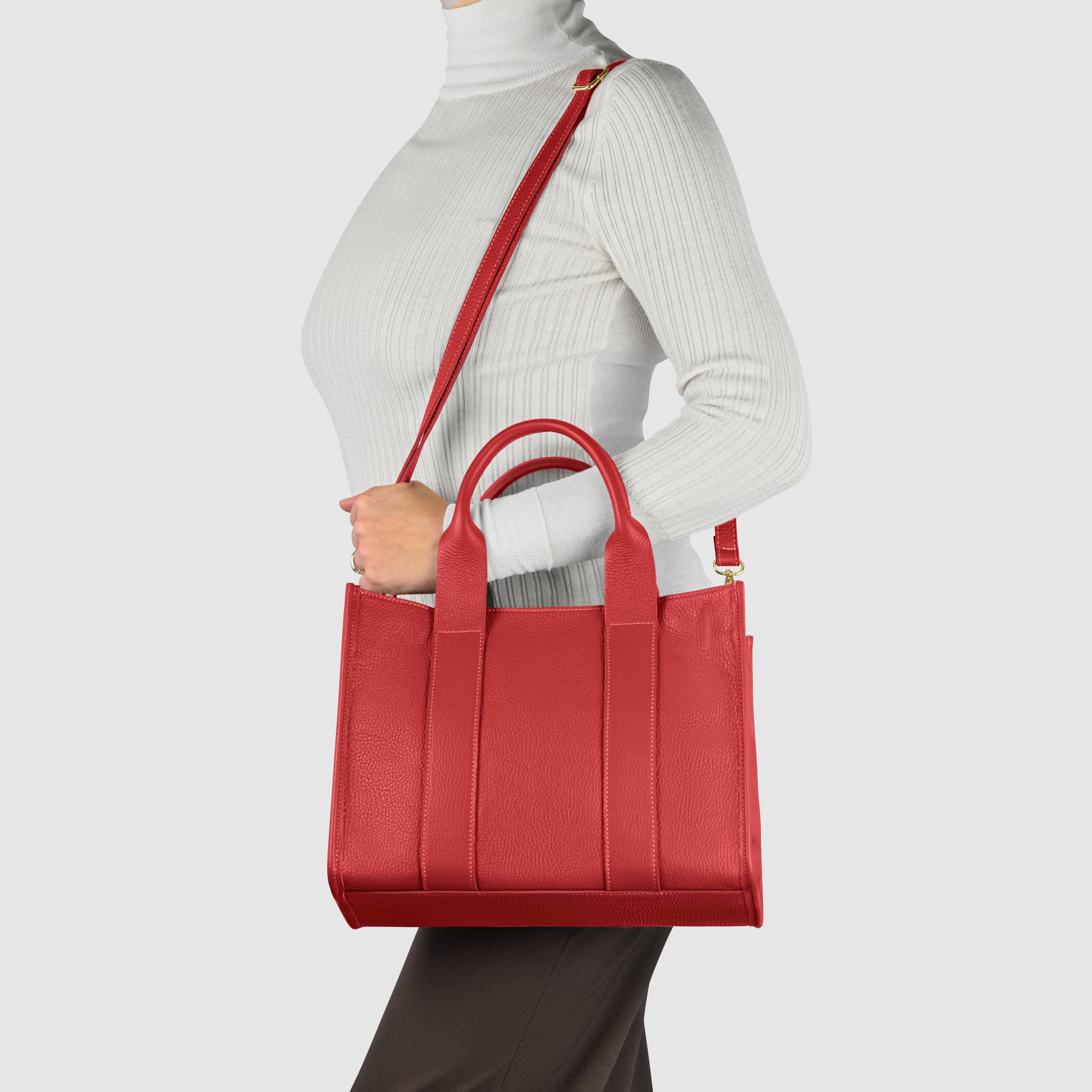 A person wearing a white turtleneck and dark pants carries a structured Italian leather handbag with both top handles and a shoulder strap against a plain light background.