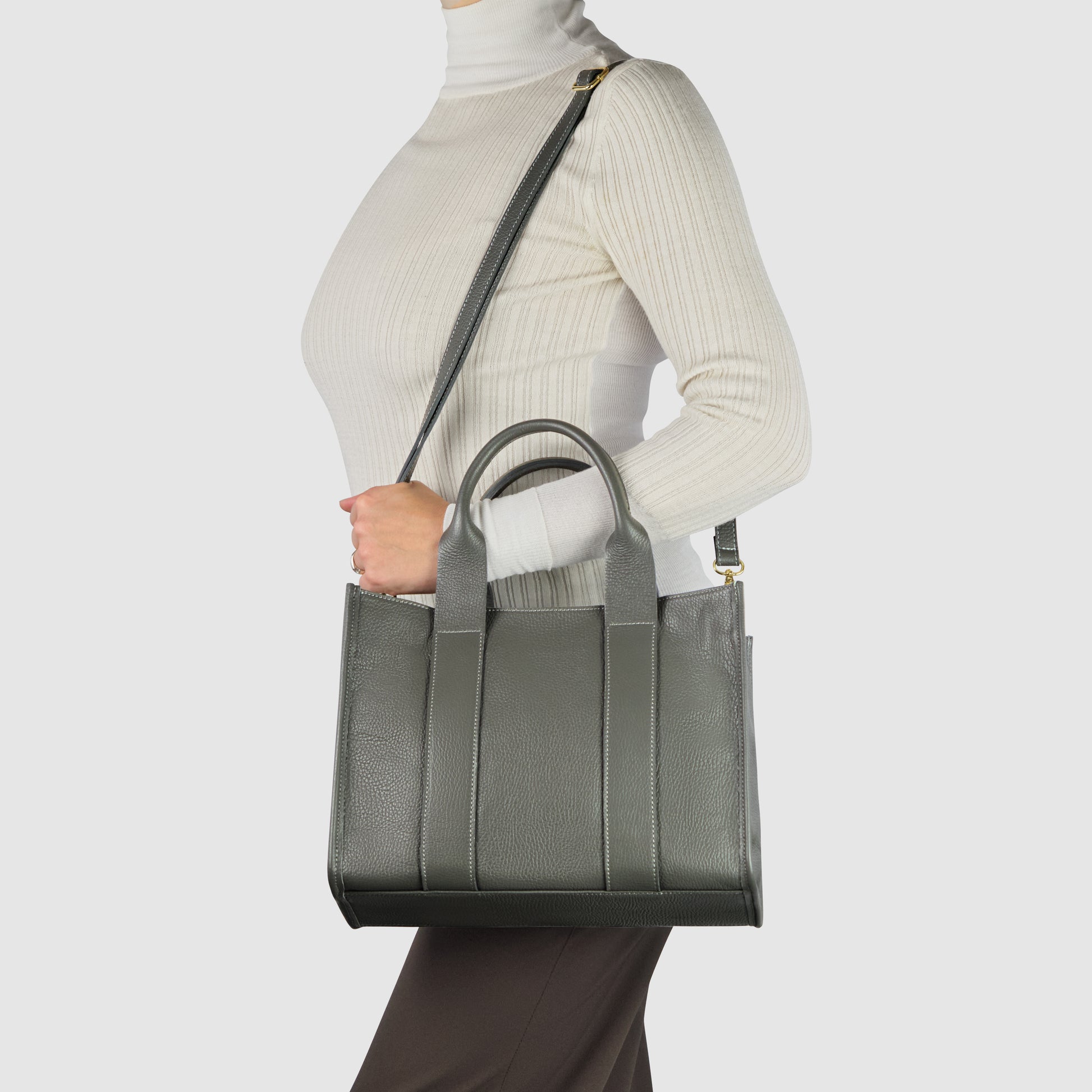 A person wearing a cream turtleneck and dark pants carries a structured Italian leather handbag with top handles and a shoulder strap against a plain light background.