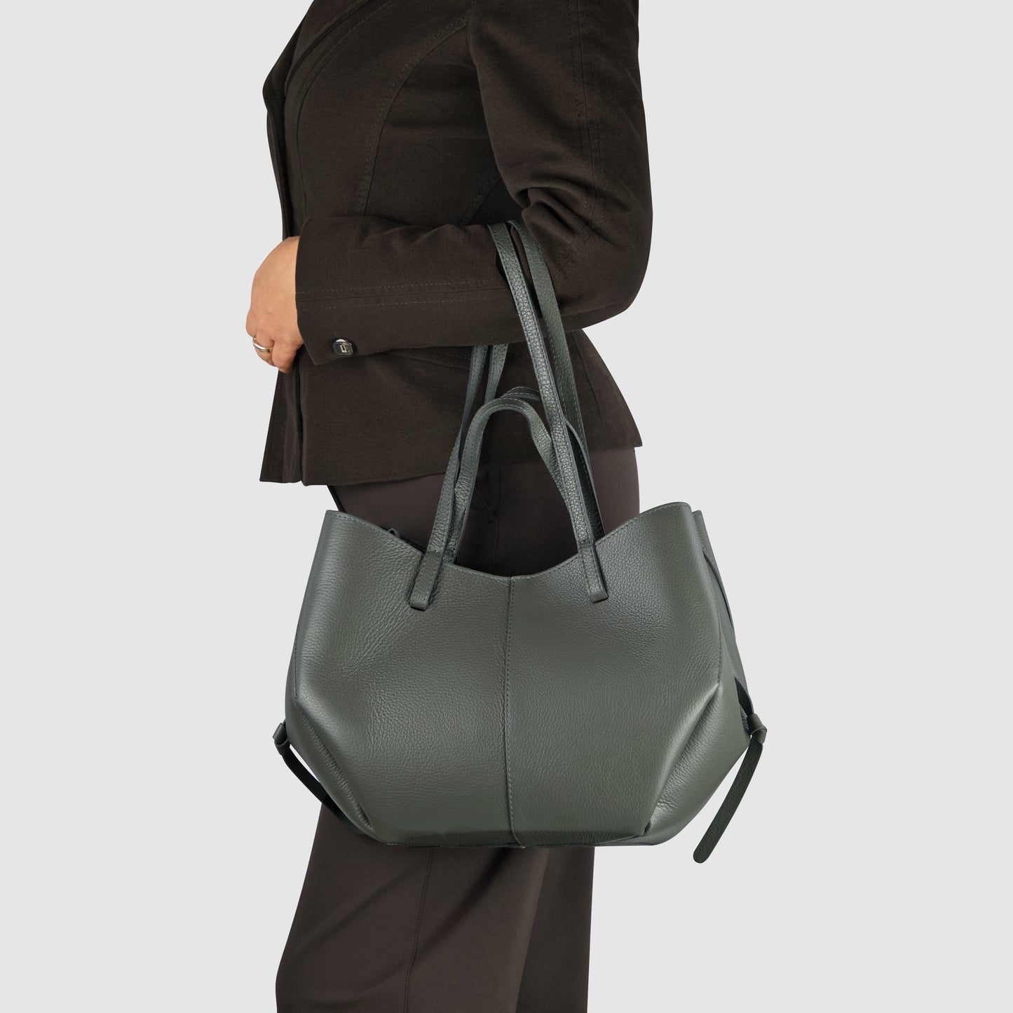 A person dressed in a dark blazer and pants holds a large, handcrafted in Italy dark green leather tote bag with double handles over their shoulder against a plain light background.