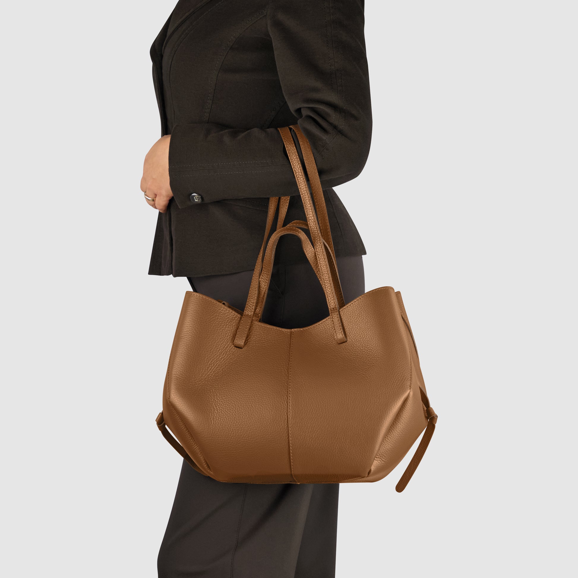 A person in a dark suit holds a large Italian leather handbag over their shoulder. The image is set against a plain light gray background, highlighting the craftsmanship and elegance of this accessory handcrafted in Italy.
