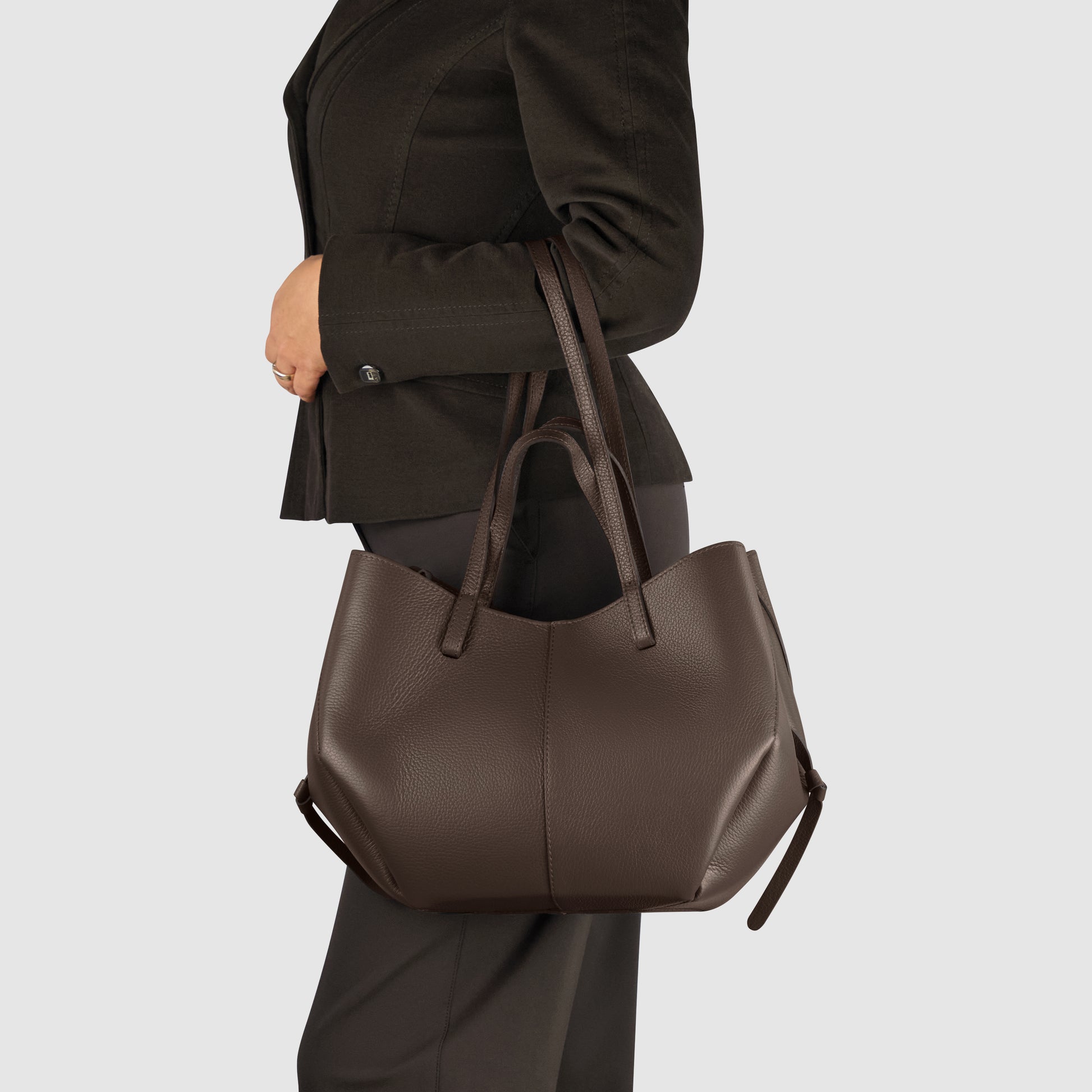 A person wearing a dark suit holds a large Italian leather handbag with double handles over their shoulder against a plain light background.