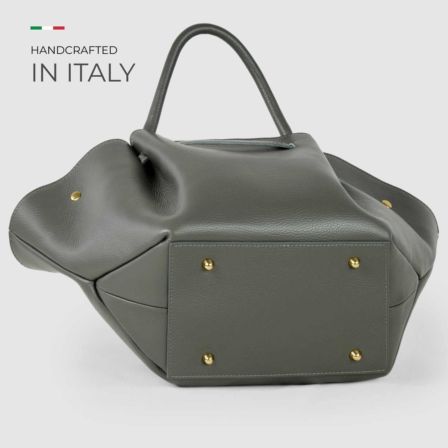 A structured, gray Italian leather bag with gold metal feet is displayed on its side. Text reads “Handcrafted in Italy” with a small Italian flag above it, all on a light gray background, showcasing true Florentine craftsmanship.