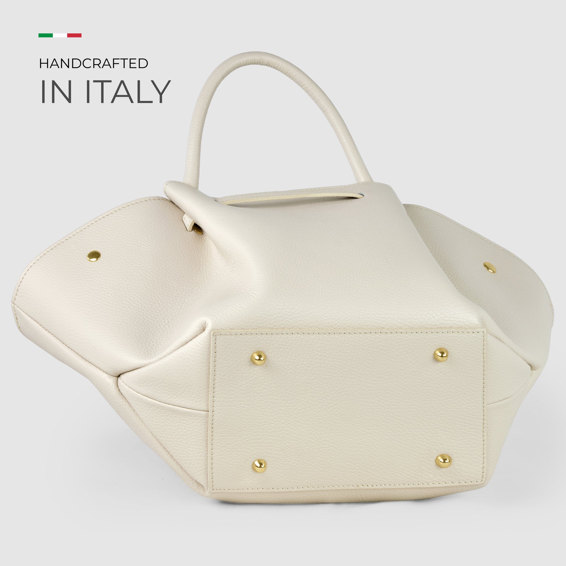 A cream-colored, uniquely structured Italian leather bag with gold studs on the bottom and two handles. Showcasing Florentine craftsmanship, "Handcrafted in Italy" and an Italian flag appear in the top left corner.