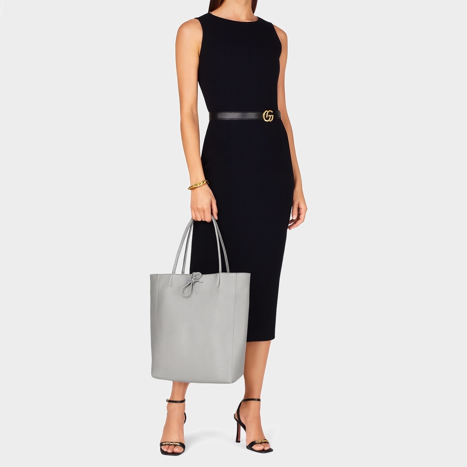 A woman in a sleeveless black midi dress, black belt, and heels stands holding a large Italian leather tote bag in light gray. She wears a gold bracelet and her hair is out of frame. The background is plain white.