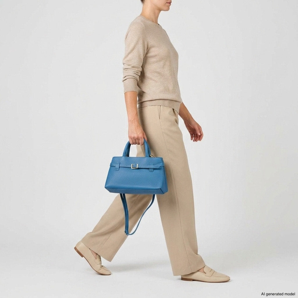 A person wearing a beige sweater and pants walks while carrying a bright blue Italian leather handbag. The background is plain and light-colored.