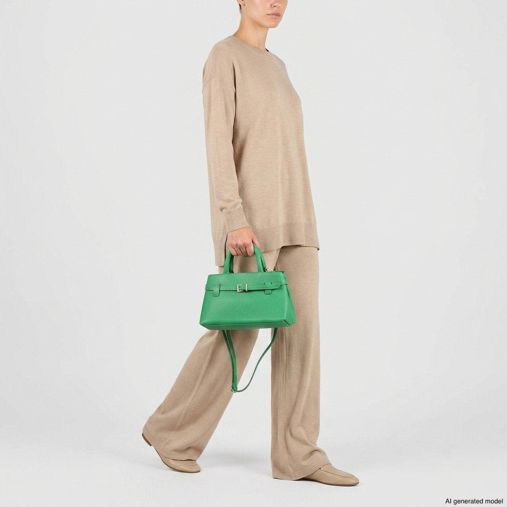Person in beige sweater and wide-leg pants holding a minimalist structured purse in green, walking against a plain white background. Face is partially out of frame.