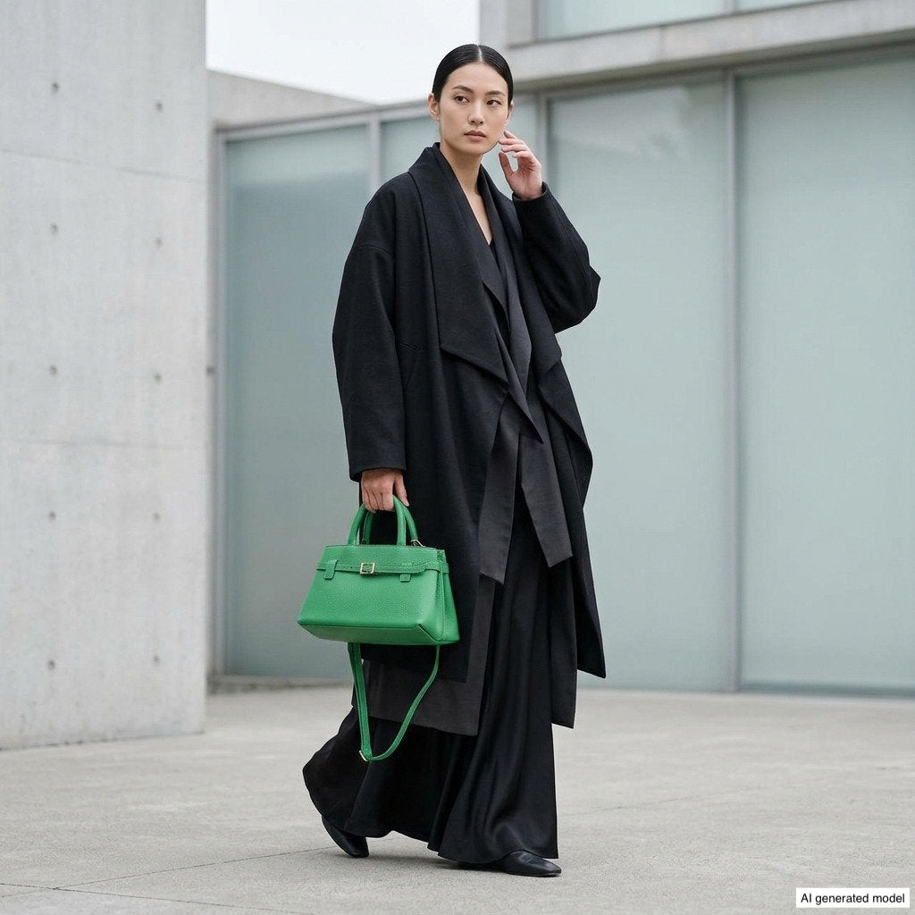 A woman in a long, black overcoat and dress stands outside a modern building, holding a bright green Italian leather handbag. She has dark hair pulled back and wears black shoes. The setting appears to be urban and minimalist.