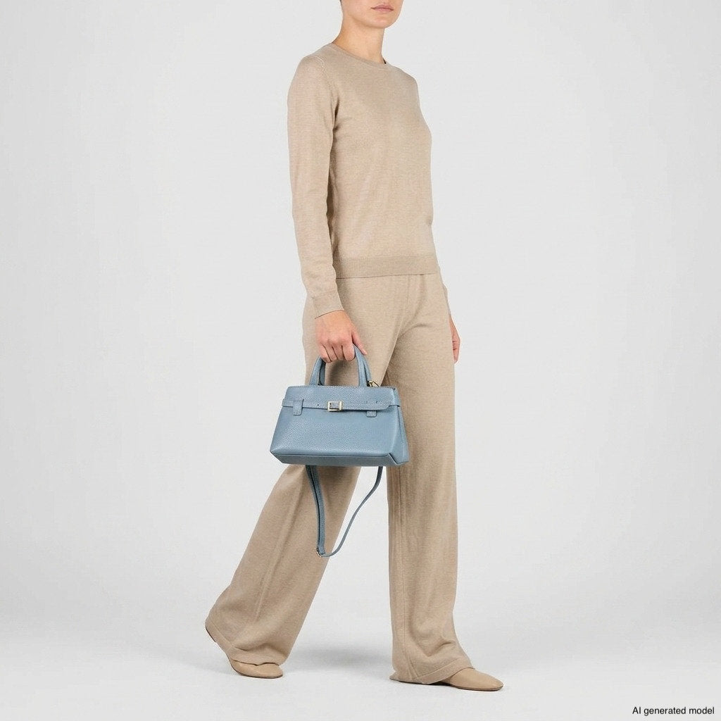 Person in a beige long-sleeve top and matching pants stands holding a minimalist structured purse in striking blue. The background is plain white. Face is partially out of frame.