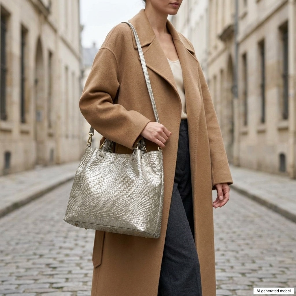 A woman wearing a camel coat and gray pants stands on a cobblestone street, holding a large metallic suede handbag. Her face is partially out of frame.