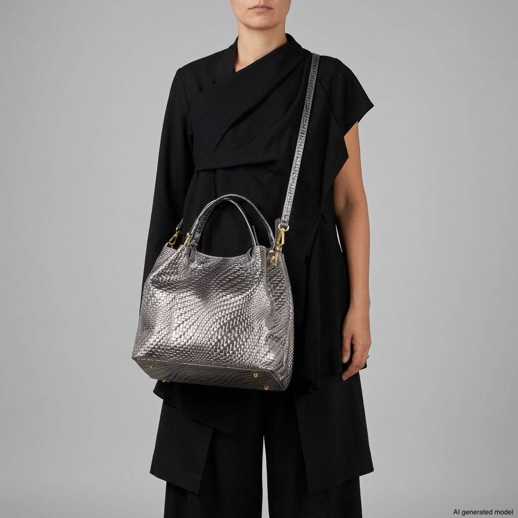 A person wearing a black asymmetrical outfit holds a large metallic suede handbag with both handles and a shoulder strap, standing against a plain gray background.