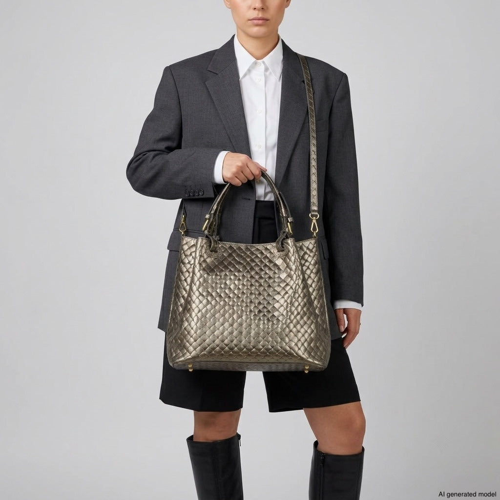 Person wearing a dark blazer, white shirt, black shorts, and black boots holds a large woven-effect handbag with both short handles and a shoulder strap. This piece highlights authentic Italian craftsmanship with its striking metallic finish. Plain gray background.