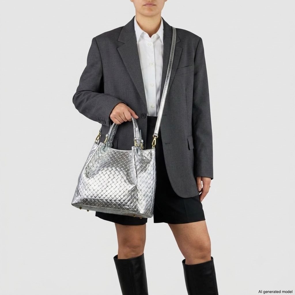Person wearing a dark blazer, white shirt, black shorts, and knee-high boots, holding a woven-effect handbag with a shiny silver finish and shoulder strap that highlights authentic Italian craftsmanship, standing against a plain light background.