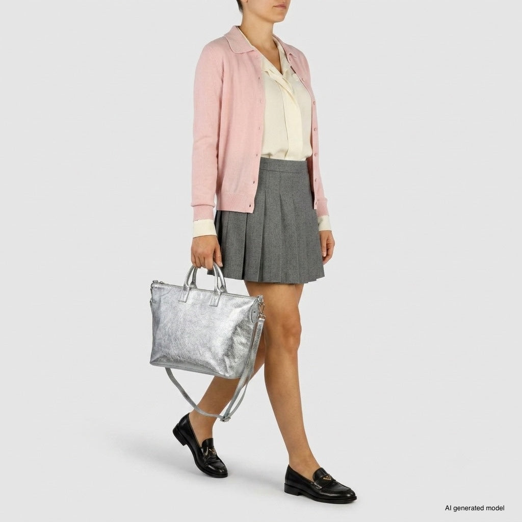 A woman wearing a light pink cardigan, cream blouse, grey pleated skirt, and black loafers holds a handcrafted Italian handbag with a metallic finish against a plain light background.