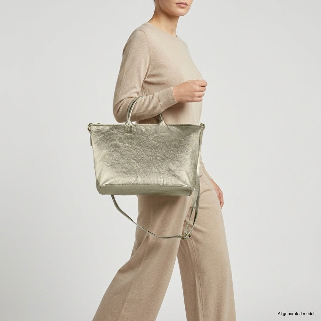 A woman in a beige long-sleeve top and wide-leg pants carries a handcrafted Italian handbag with a metallic finish, featuring both short handles and a long strap, as she walks against a plain light background.