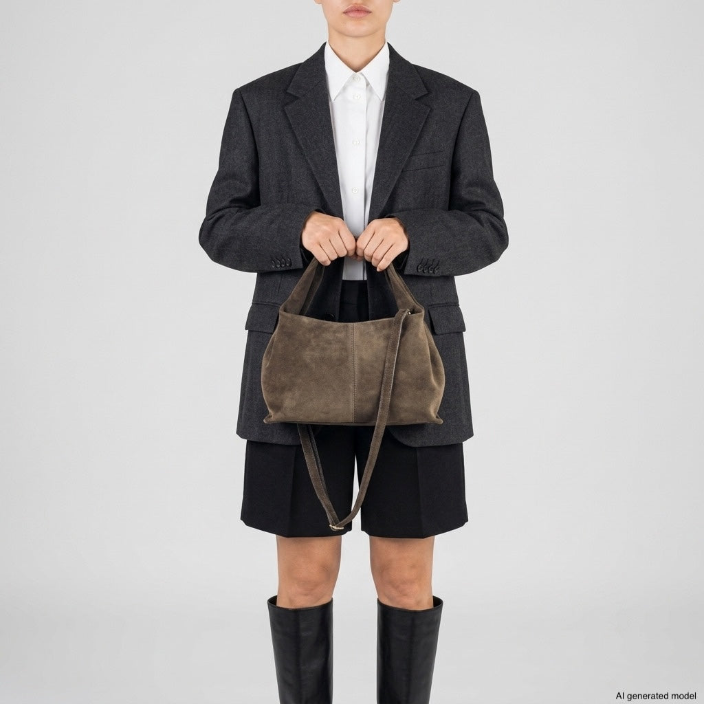 A person wearing a dark blazer, white shirt, black shorts, and knee-high black boots is holding a brown suede handbag showcasing Italian craftsmanship with both hands against a plain light background.